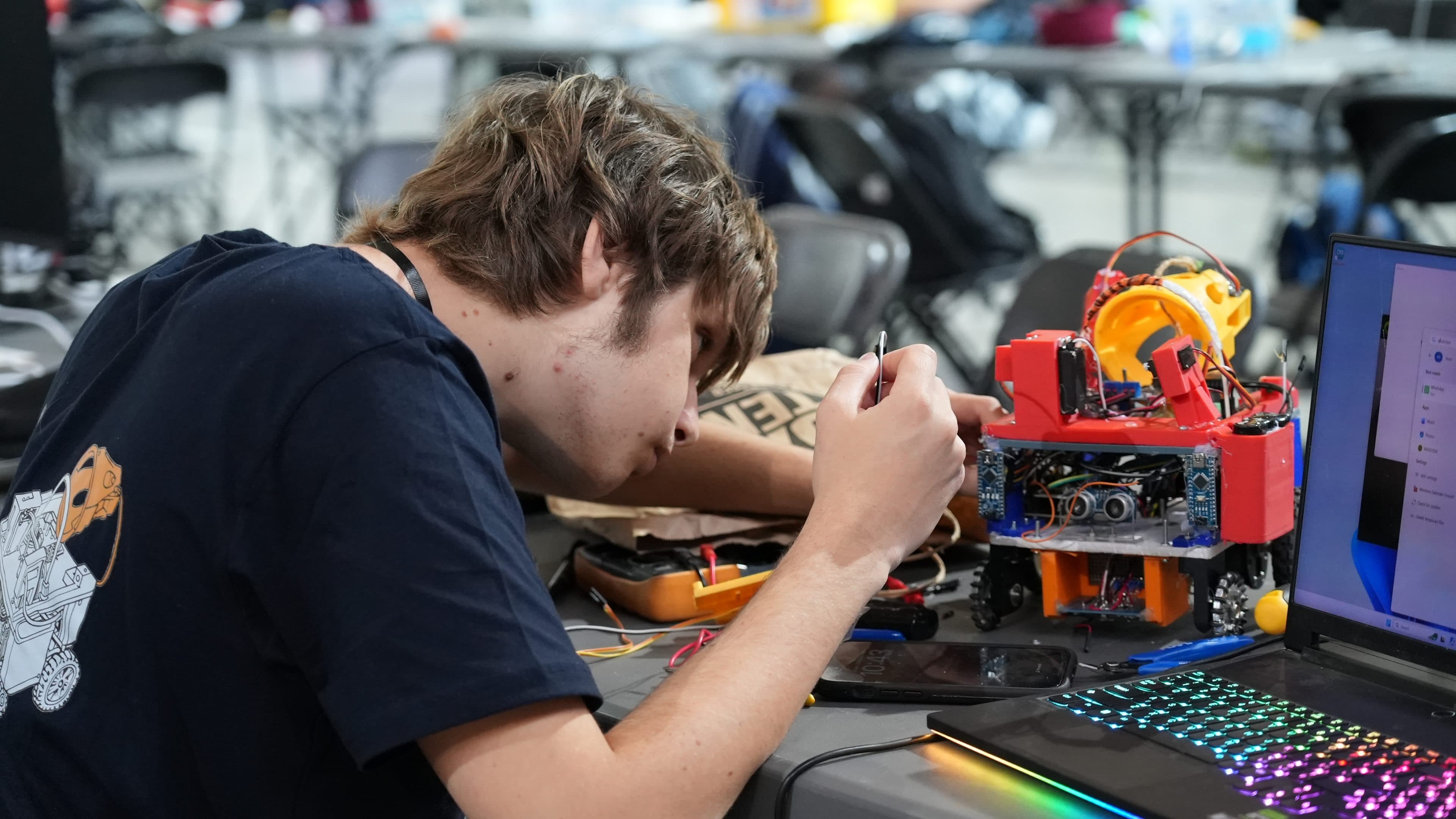 Ein junger Mensch arbeitet konzentriert daran, einen kleinen rot-gelben Roboter an einem Tisch zusammenzubauen oder zu reparieren, umgeben von elektronischem Werkzeug, Kabeln, einem Laptop mit bunter Tastatur und weiterer Ausrüstung.