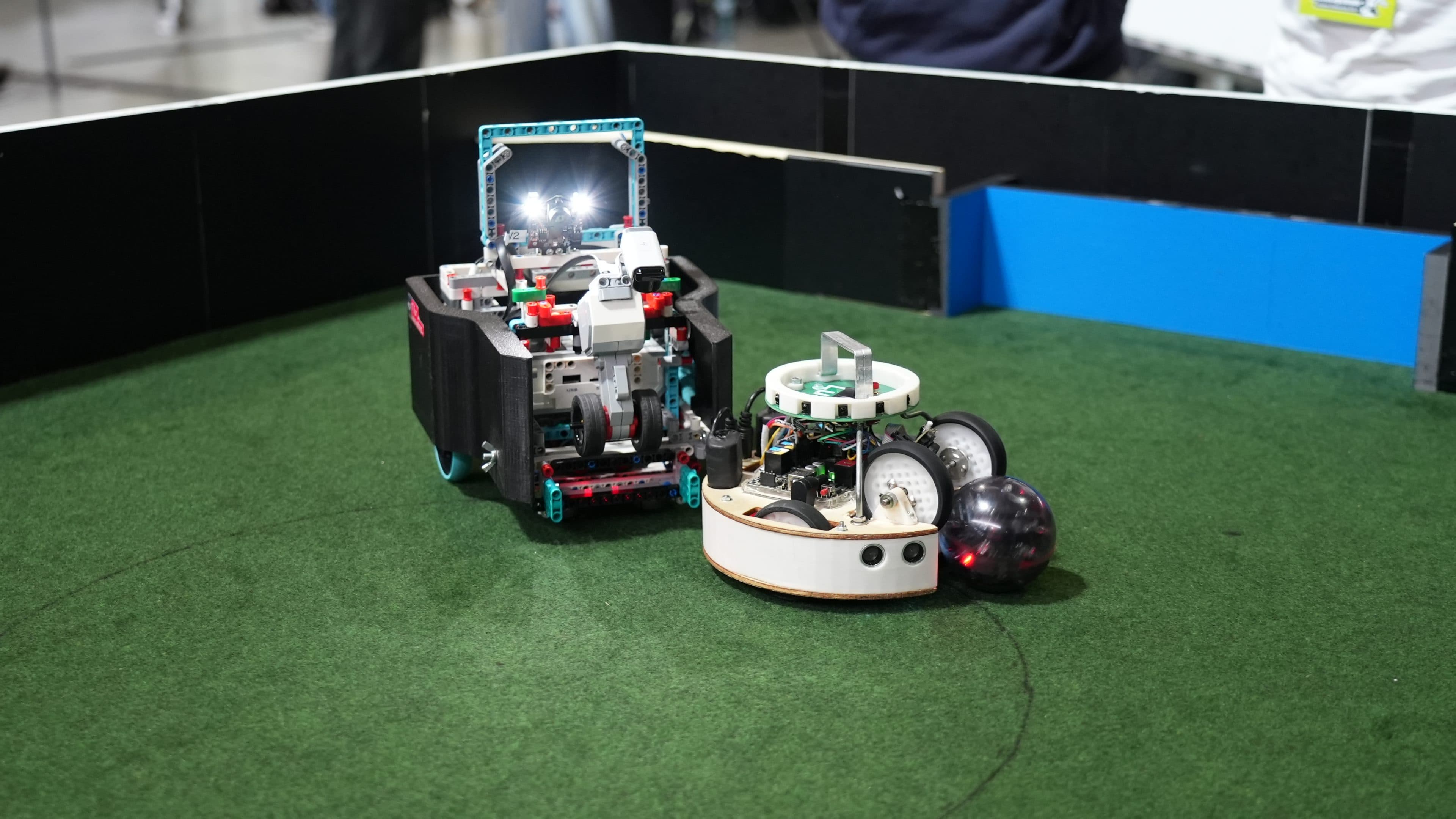 Zwei kleine Roboter auf einer grünen Kunstrasenfläche, die scheinbar um einen schwarzen Ball wetteifern oder mit ihm interagieren. Die Roboter haben freiliegende Schaltkreise und Räder, im Hintergrund ist eine schwarze Barriere zu sehen.