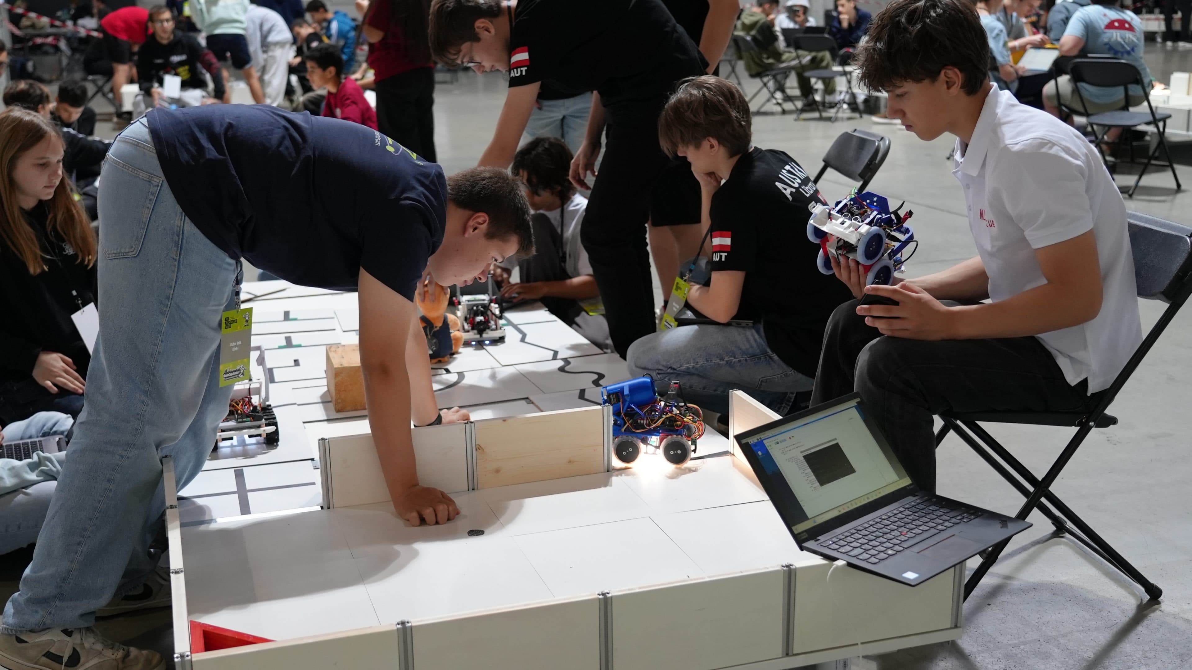 Mehrere Jugendliche nehmen an einem Robotikwettbewerb teil und nehmen auf einer markierten Arena die Feinjustierung und das Testen kleiner Radroboter vor, während andere zuschauen und an ihren Laptops arbeiten. Der Schauplatz scheint ein großer Veranstaltungsraum zu sein.