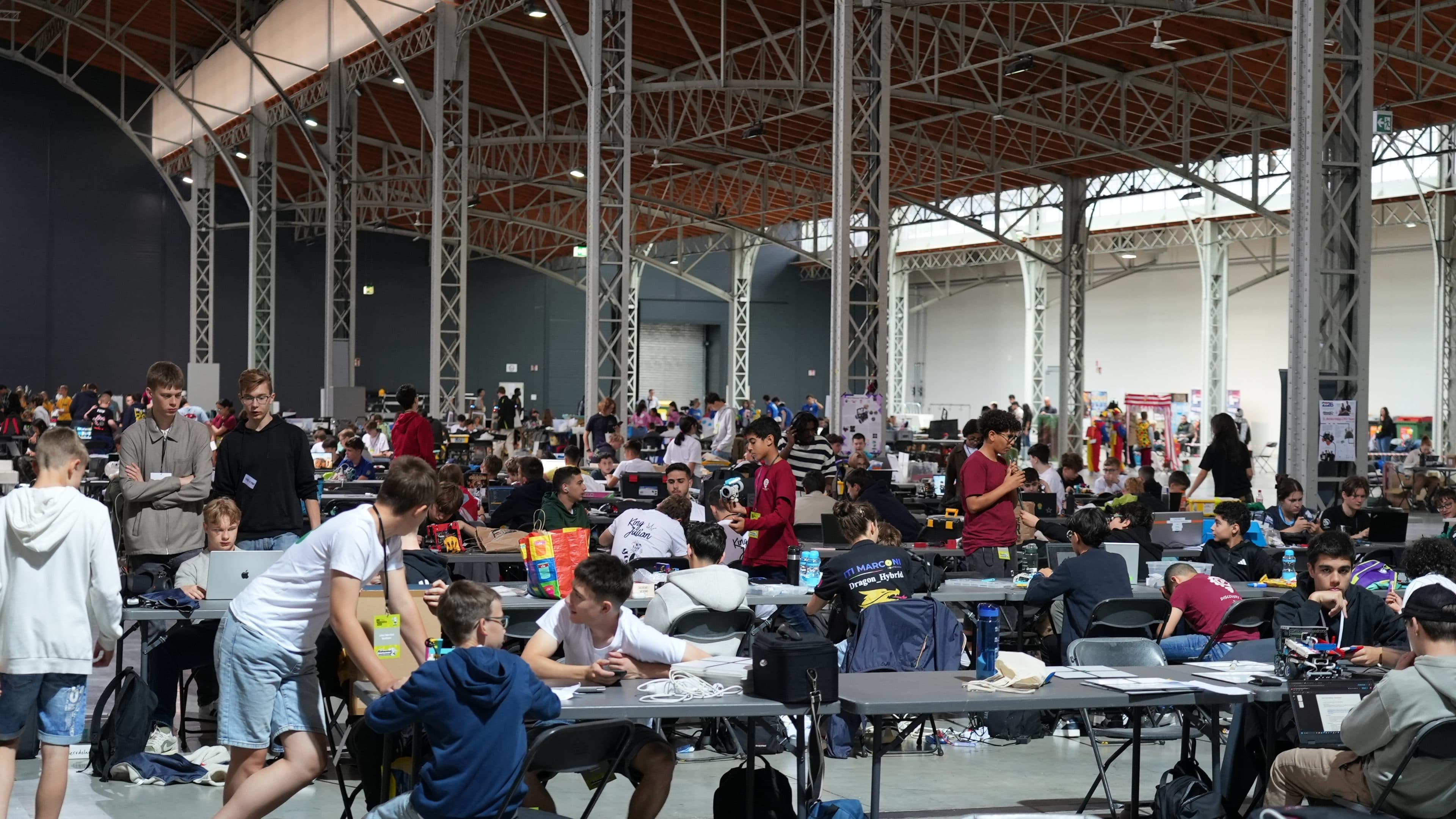 Ein großer Saal, gefüllt mit vielen jungen Menschen, die in Gruppen an Tischen arbeiten, auf denen Laptops, Papiere und elektronische Geräte liegen – ein Bild, das an einen geschäftigen Hackathon oder eine gemeinschaftliche Veranstaltung unter einer hohen Decke mit Stahlträgerwerk erinnert.