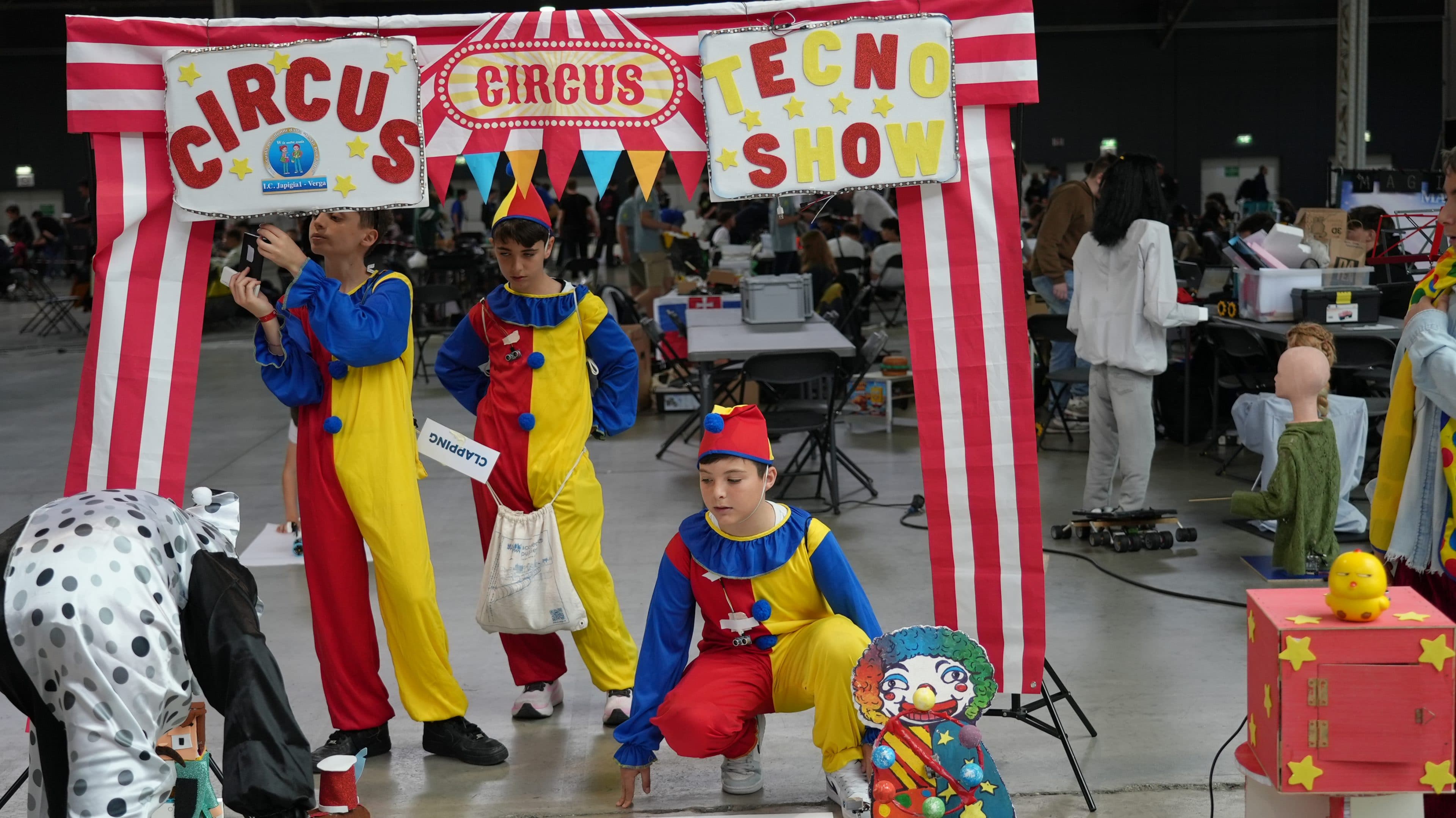 Drei als Clowns verkleidete Personen stehen bei einer Veranstaltung in einer Halle unter einem bunten Schild mit der Aufschrift „Circus Tecno Show“, im Hintergrund sind Tische, Ausrüstung und Besucher zu sehen. Auf dem Boden in der Nähe liegen eine Clownpuppe und Requisiten.