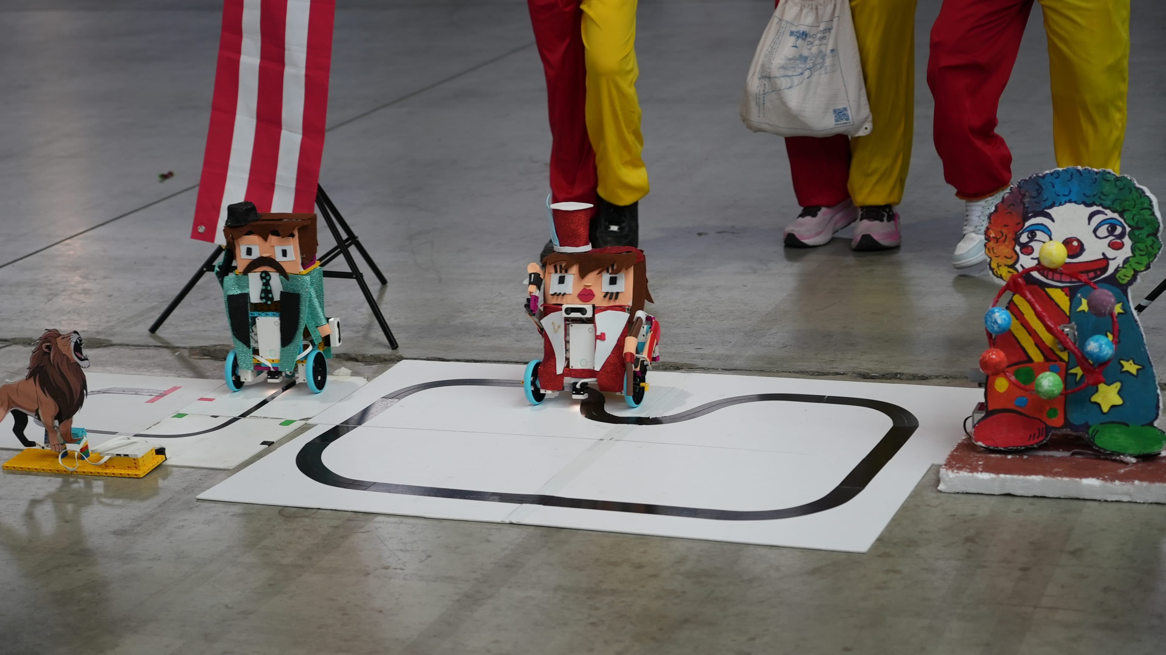 Zwei kleine Roboter auf Rädern im Clown-Design stehen am Anfang einer Bahn auf dem Boden, während hinter ihnen Menschen in bunten Kostümen und eine große Clownfigur stehen. Links ist teilweise eine amerikanische Flagge zu sehen.