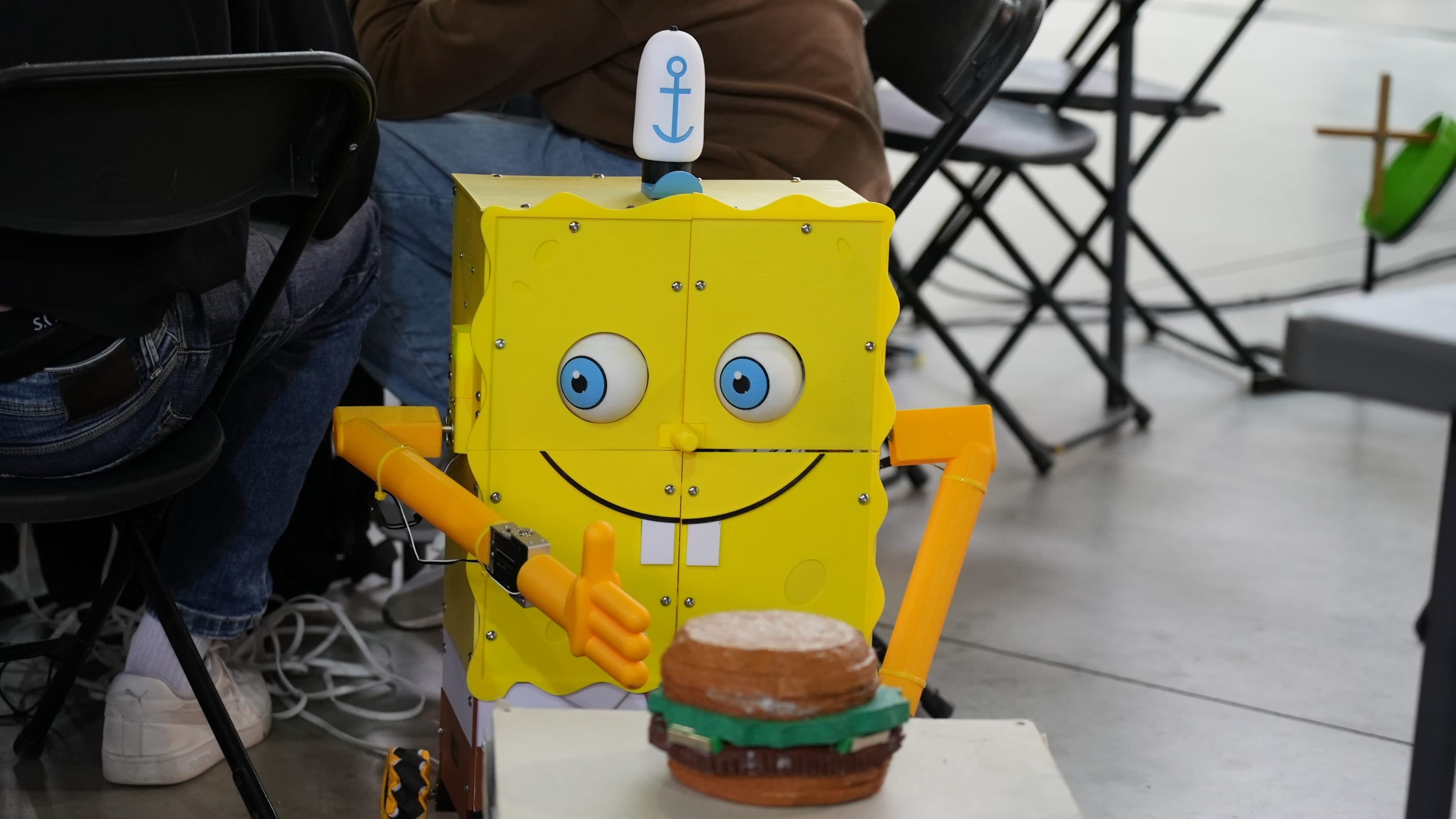 Ein Roboter, der wie SpongeBob Schwammkopf gestaltet ist – mit einem lächelnden Gesicht und einem blauen Anker auf dem Kopf –, sitzt zwischen schwarzen Stühlen und blickt auf einen Spielzeug-Hamburger auf einem Tisch.