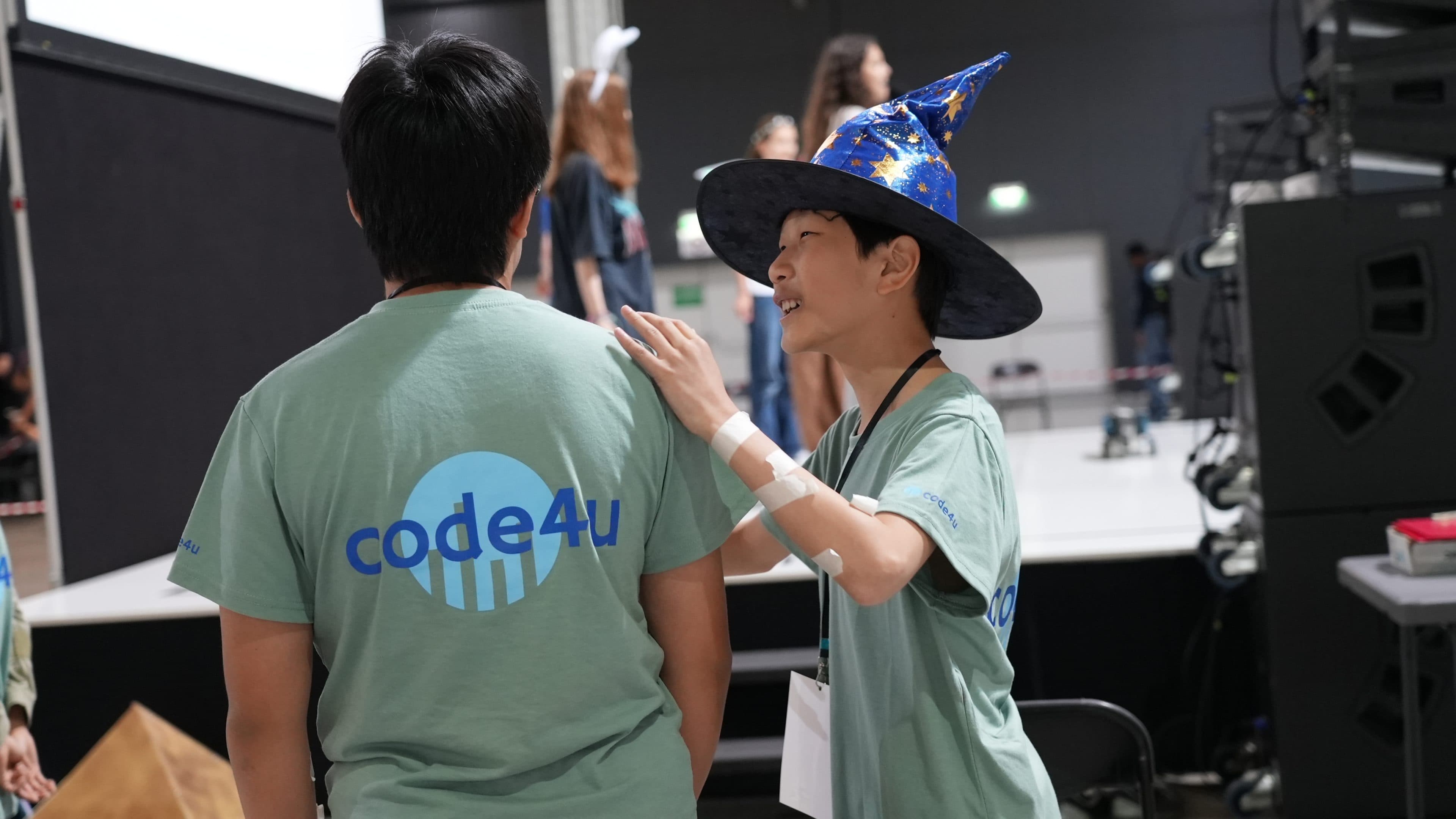 Ein lächelnder Junge mit einem blauen Zaubererhut berührt die Schulter eines anderen Jungen. Beide tragen hellgrüne „code4u“-T-Shirts. Sie stehen in der Nähe einer Bühne, im Hintergrund sind Menschen und Technik zu sehen.