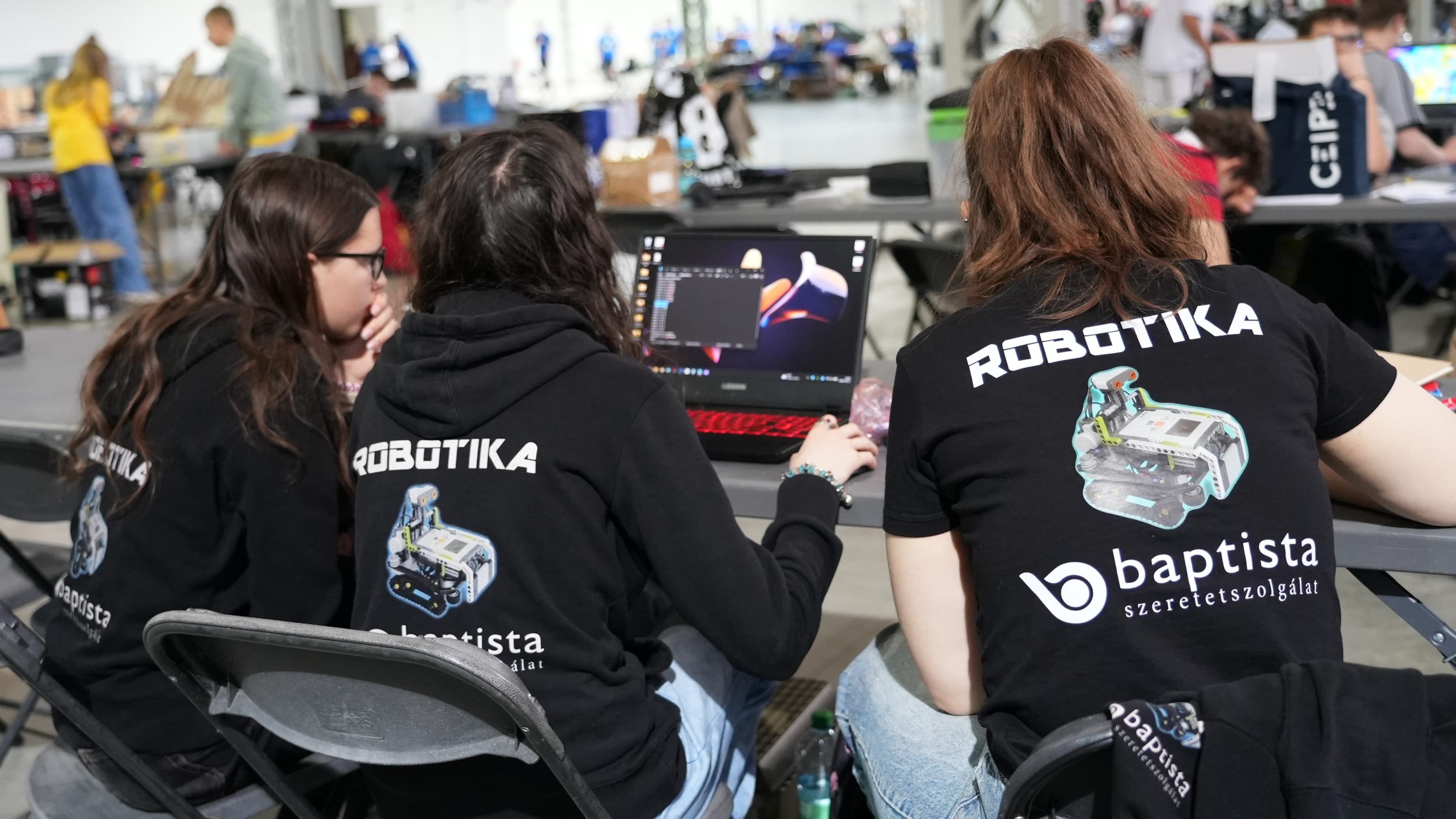 Drei Personen, die „ROBOTIKA“-T-Shirts tragen, sitzen an einem Tisch und arbeiten an einem Laptop. Sie scheinen sich auf einer Robotik-Veranstaltung oder einem Wettbewerb zu befinden, da im Hintergrund andere Teams und Ausrüstung zu sehen sind.