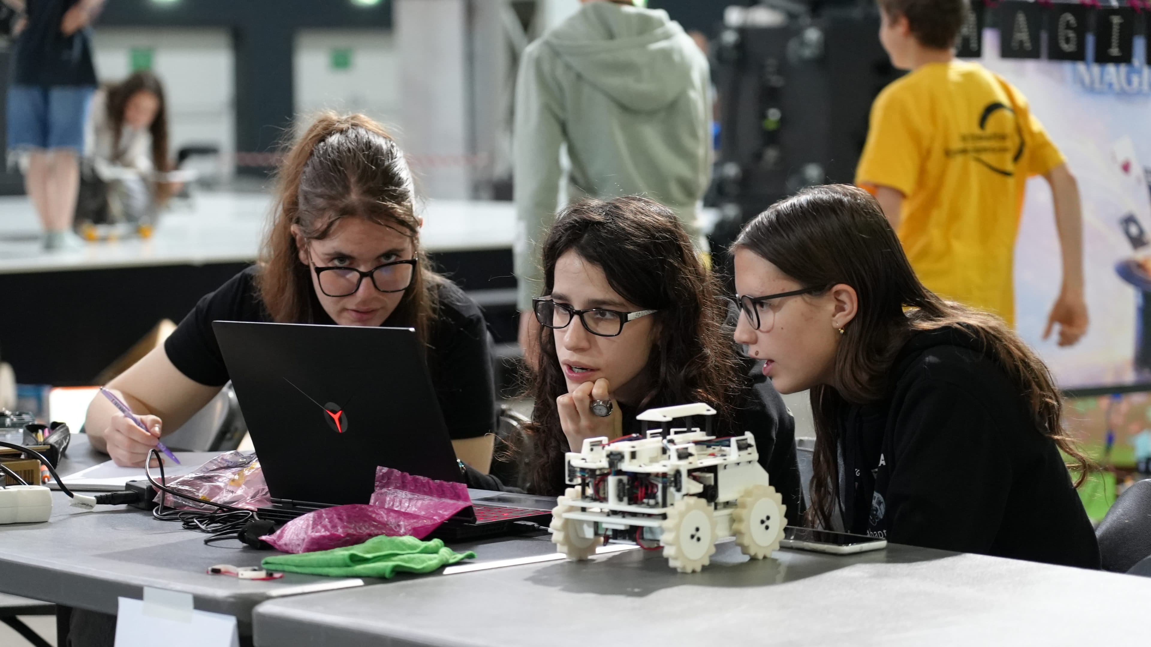 Drei junge Frauen mit Brille arbeiten konzentriert an einem Laptop, auf dem Tisch stehen ein kleiner weißer Roboter und elektronische Bauteile. Sie wirken konzentriert und arbeiten eng zusammen bei einer Veranstaltung zum Thema Robotik oder MINT.