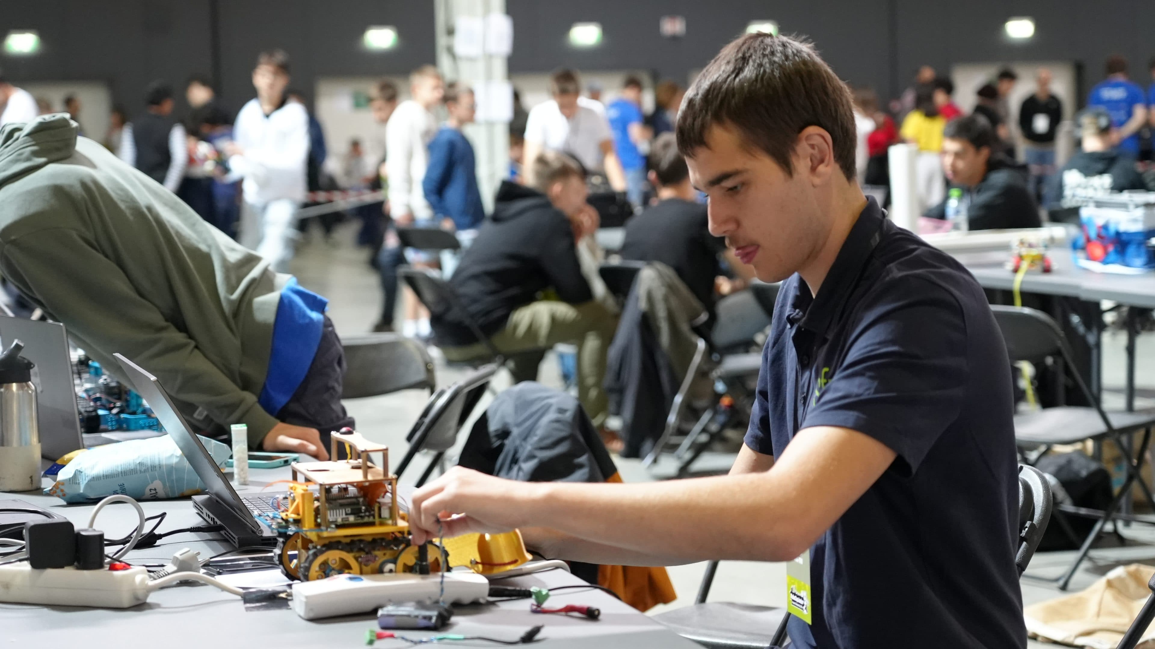 Ein junger Mann arbeitet an einem Tisch bei einer gut besuchten Indoor-Veranstaltung am Zusammenbau eines kleinen Roboterfahrzeugs; um ihn herum liegen Werkzeuge und elektronische Bauteile, im Hintergrund sind weitere Teilnehmer zu sehen.
