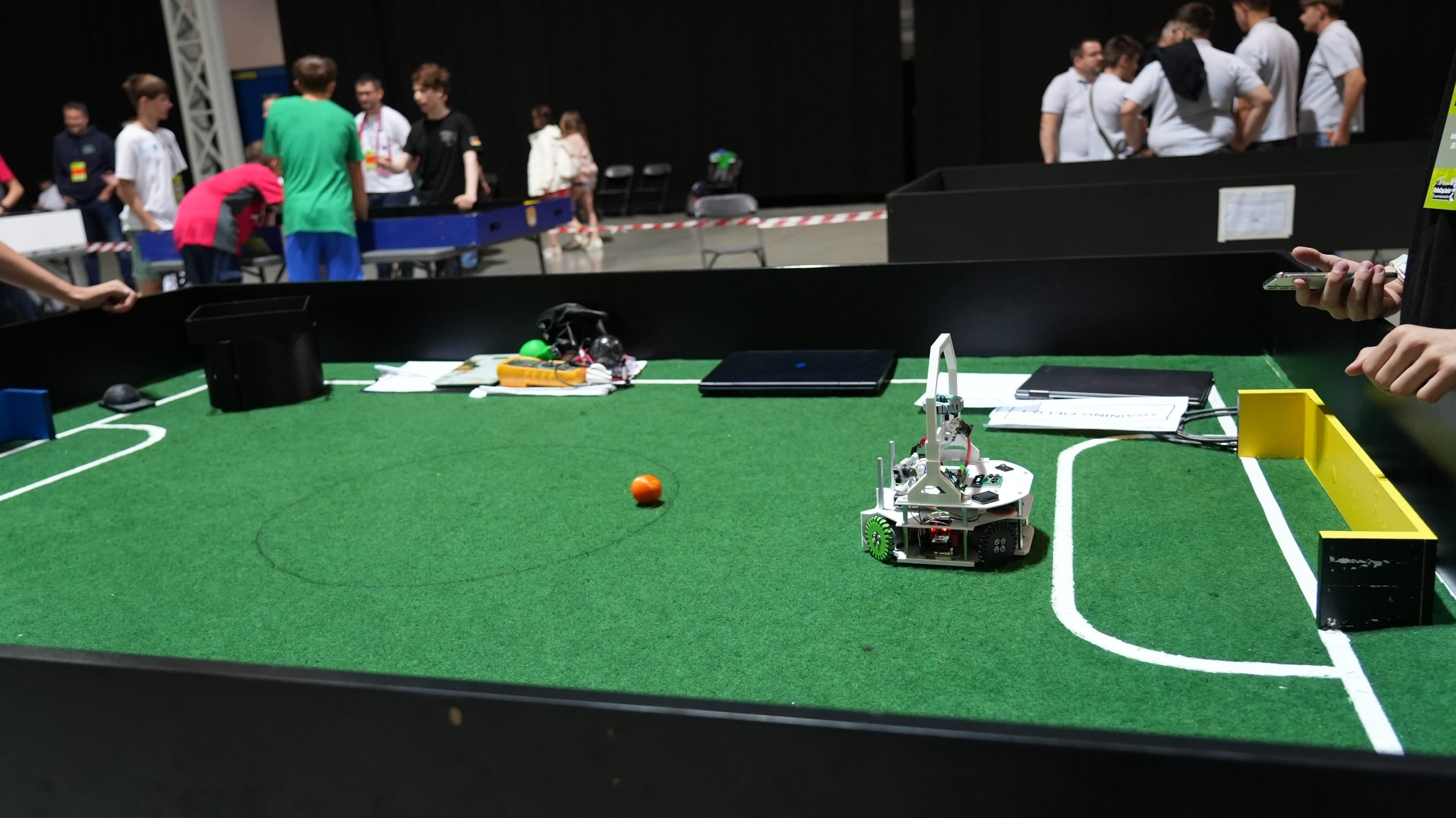 Ein kleiner weißer Roboter befindet sich auf einem grünen Kunstrasenfeld mit weißen Linien, einem orangefarbenen Ball sowie gelben und schwarzen Toren. Im Hintergrund haben sich mehrere Menschen um das Spielfeld versammelt – offenbar bei einem Robotikwettbewerb oder einer ähnlichen Veranstaltung.