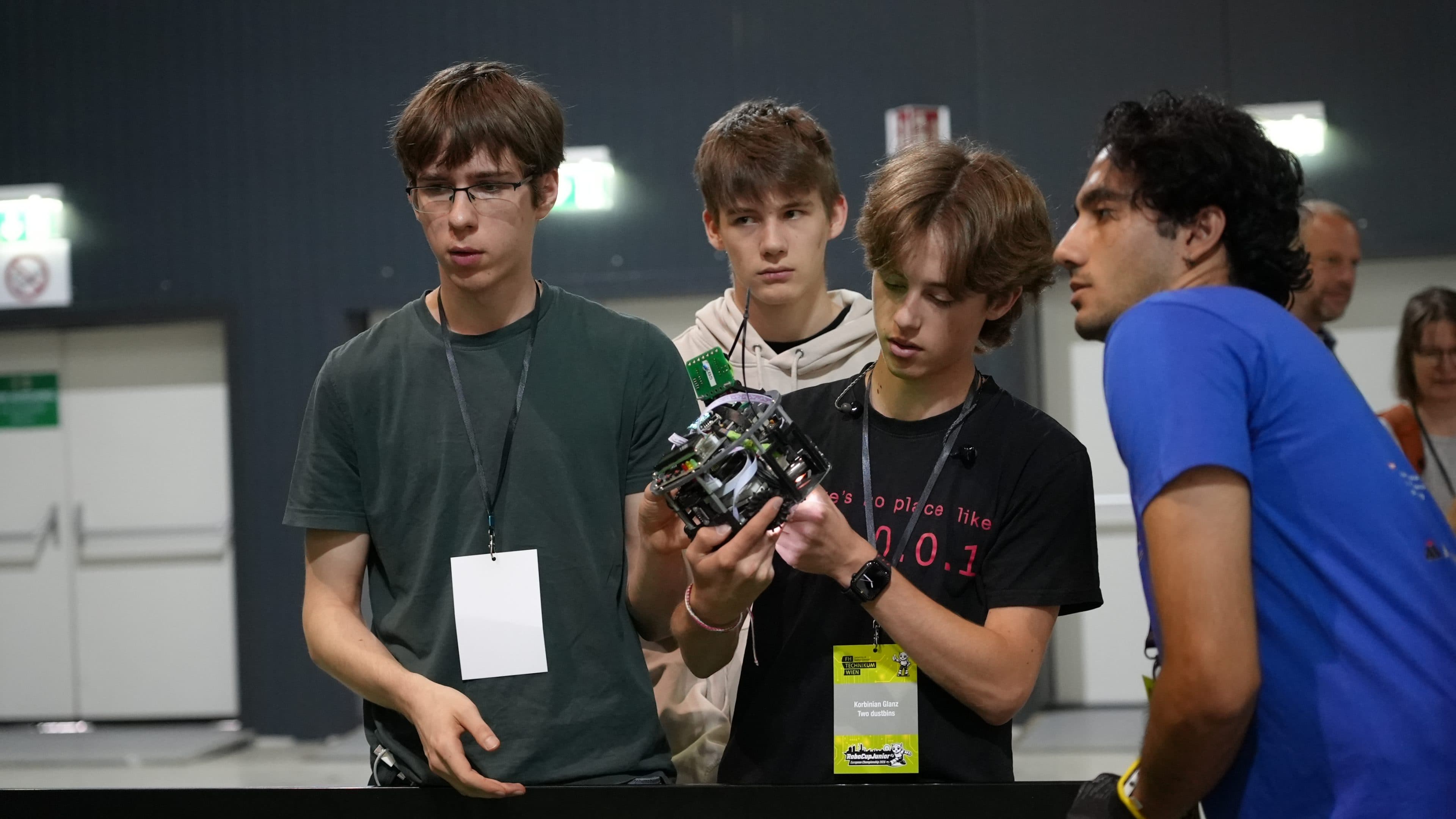 Vier junge Leute stehen zusammen in einem Raum und richten ihre Aufmerksamkeit auf einen kleinen Roboter, den einer von ihnen in der Hand hält. Sie tragen Namensschilder und Schlüsselbänder, was darauf hindeutet, dass sie an einem Wettbewerb oder einer Veranstaltung teilnehmen. Im Hintergrund beobachten zwei Erwachsene die Szene.