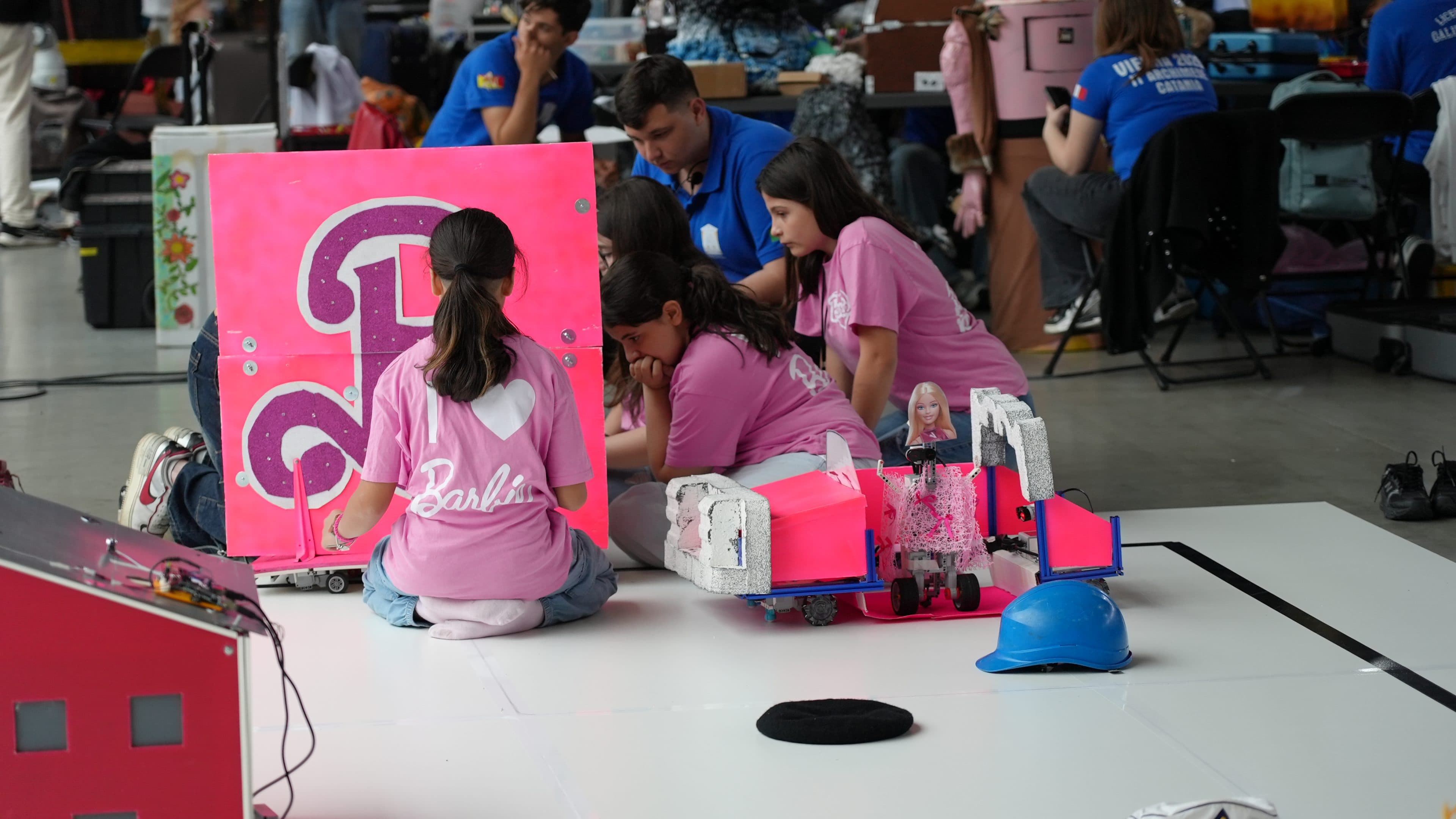 Vier Mädchen in einheitlichen rosa Barbie-Shirts arbeiten bei einer Robotik-Veranstaltung gemeinsam an einer rosa Projekttafel. Ein rosa-blauer Roboter, ein blauer Schutzhelm und weiteres Material liegen daneben auf dem Boden.