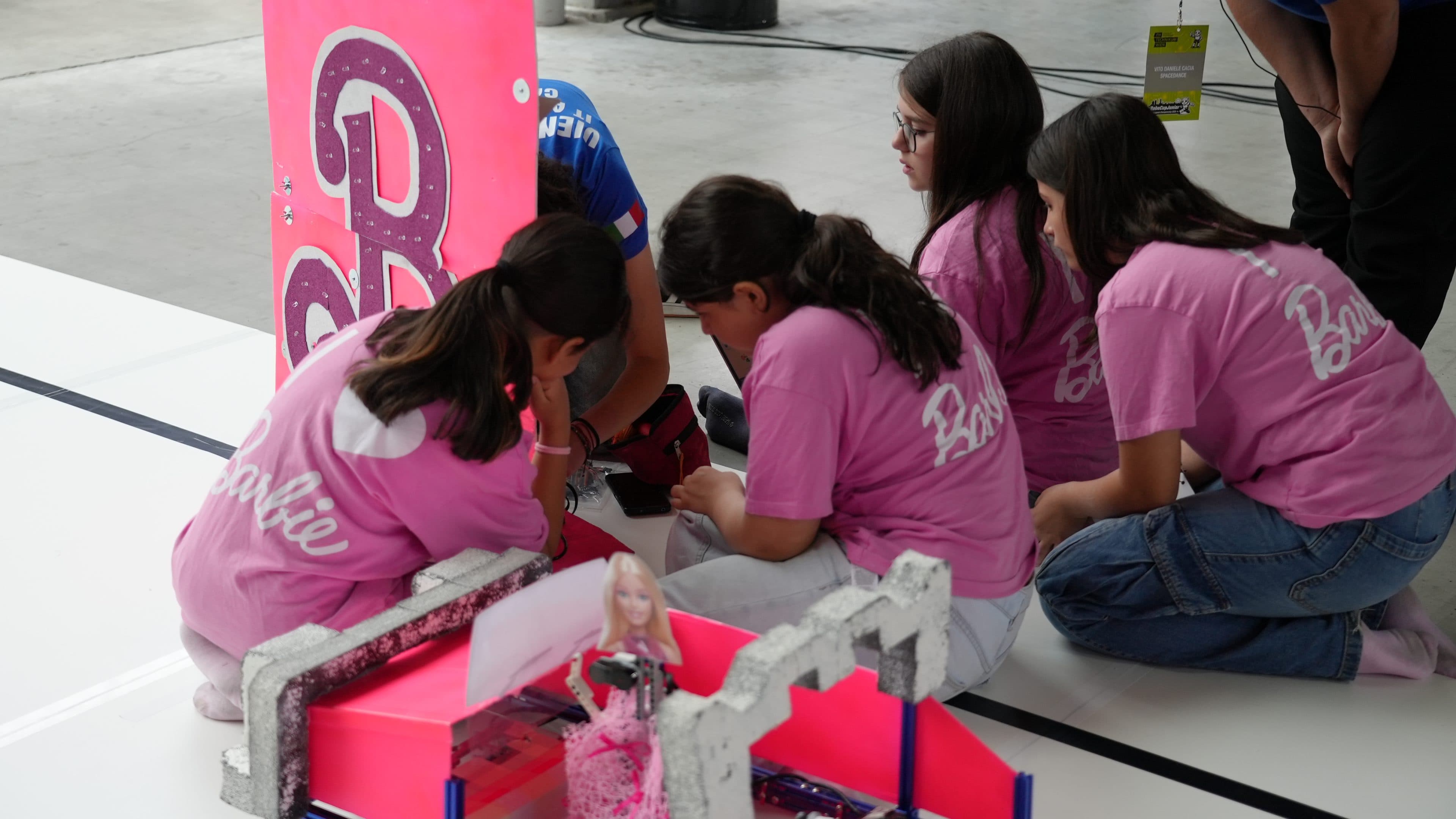 Vier Mädchen in rosa Barbie-Shirts knien auf dem Boden um ein leuchtend rosa Display herum und arbeiten gemeinsam an einem Projekt im Rahmen einer Robotik- oder MINT-Veranstaltung. Im Vordergrund sind eine Barbie-Puppe und eine Miniaturanlage zu sehen.