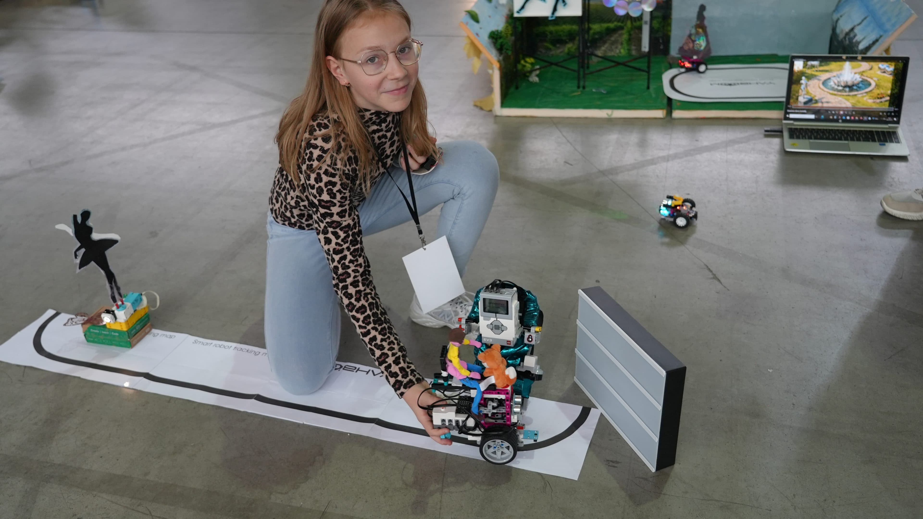 Ein Mädchen mit Brille und einem Oberteil mit Leopardenmuster kniet auf dem Boden und steuert einen bunten Roboter auf einer Papierstrecke, während im Hintergrund weitere kleine Roboter und Displays zu sehen sind.