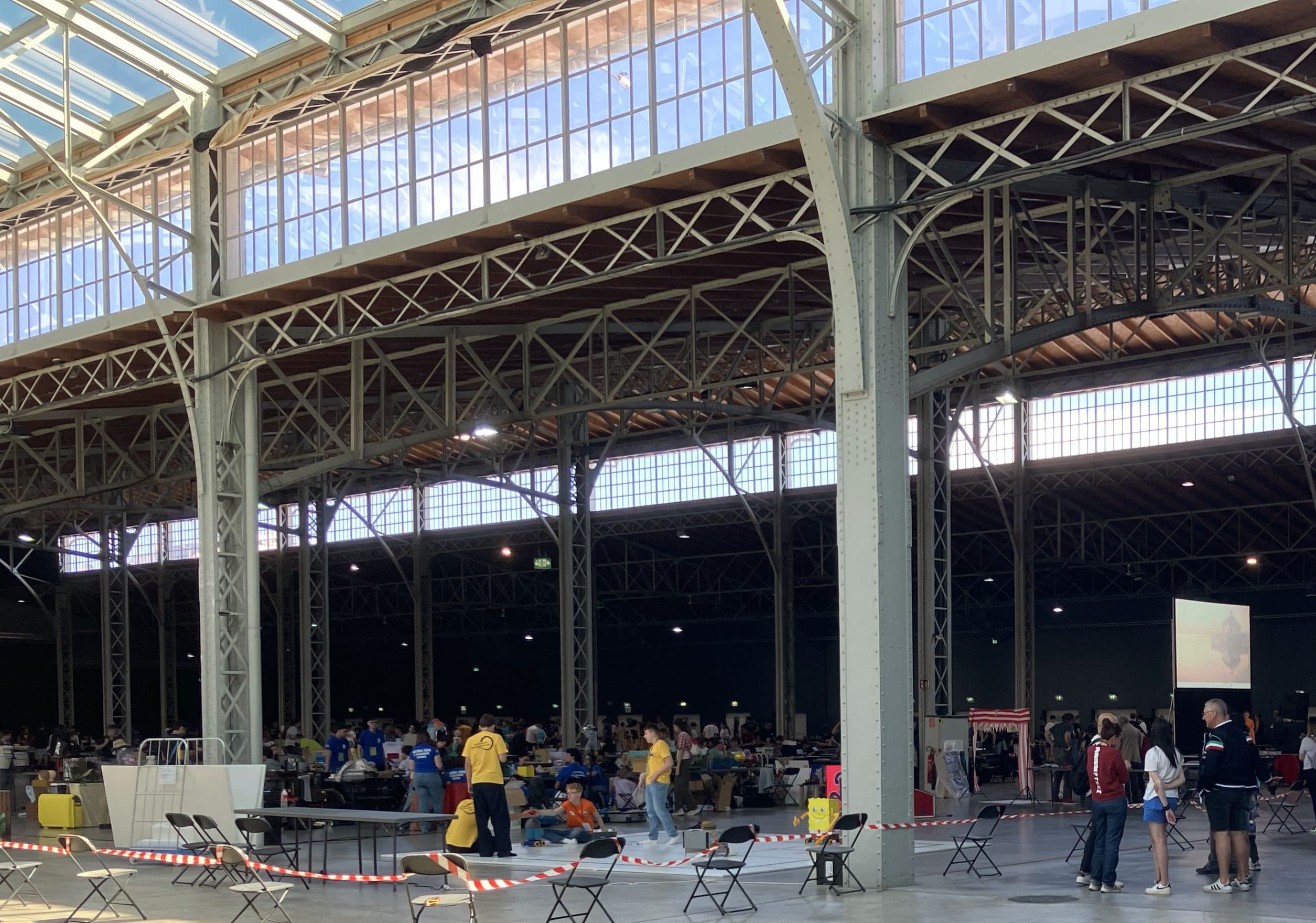 Eine große Halle mit hohen Decken, vereinzelten Personen und mehreren Tischen. Im Hintergrund arbeiten einige Gruppen an den Tischen, während ein paar Personen in der Mitte des Raums in der Nähe einer Leinwand stehen. Der Raum ist geräumig und gut beleuchtet.