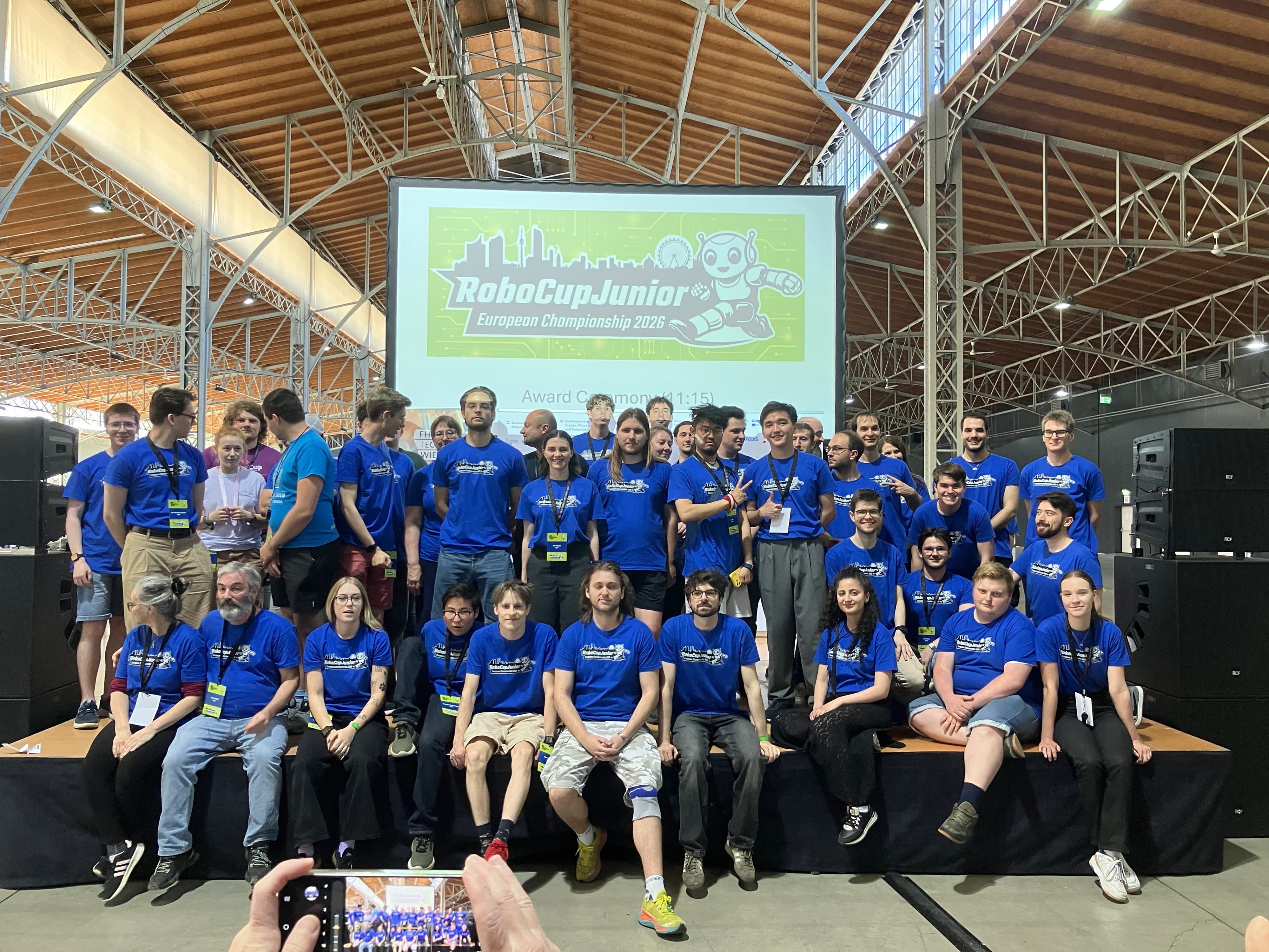 Eine große Gruppe von Menschen in einheitlichen blauen T-Shirts posiert gemeinsam auf einer Bühne vor einem Schild mit der Aufschrift „RoboCupJunior-Europameisterschaft 2023“ und lächelt für ein Foto in einer geräumigen Halle mit hohen Decken und sichtbaren Holzbalken.