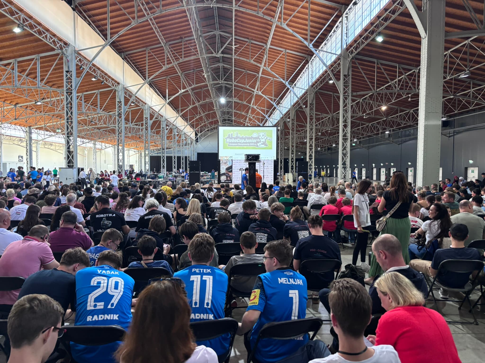 Eine große Gruppe von Menschen sitzt in Stuhlreihen in einem geräumigen Saal mit hoher Holzdecke und verfolgt eine Präsentation, die auf eine Leinwand im vorderen Bereich projiziert wird. Einige Teilnehmer tragen einheitliche Sporttrikots.