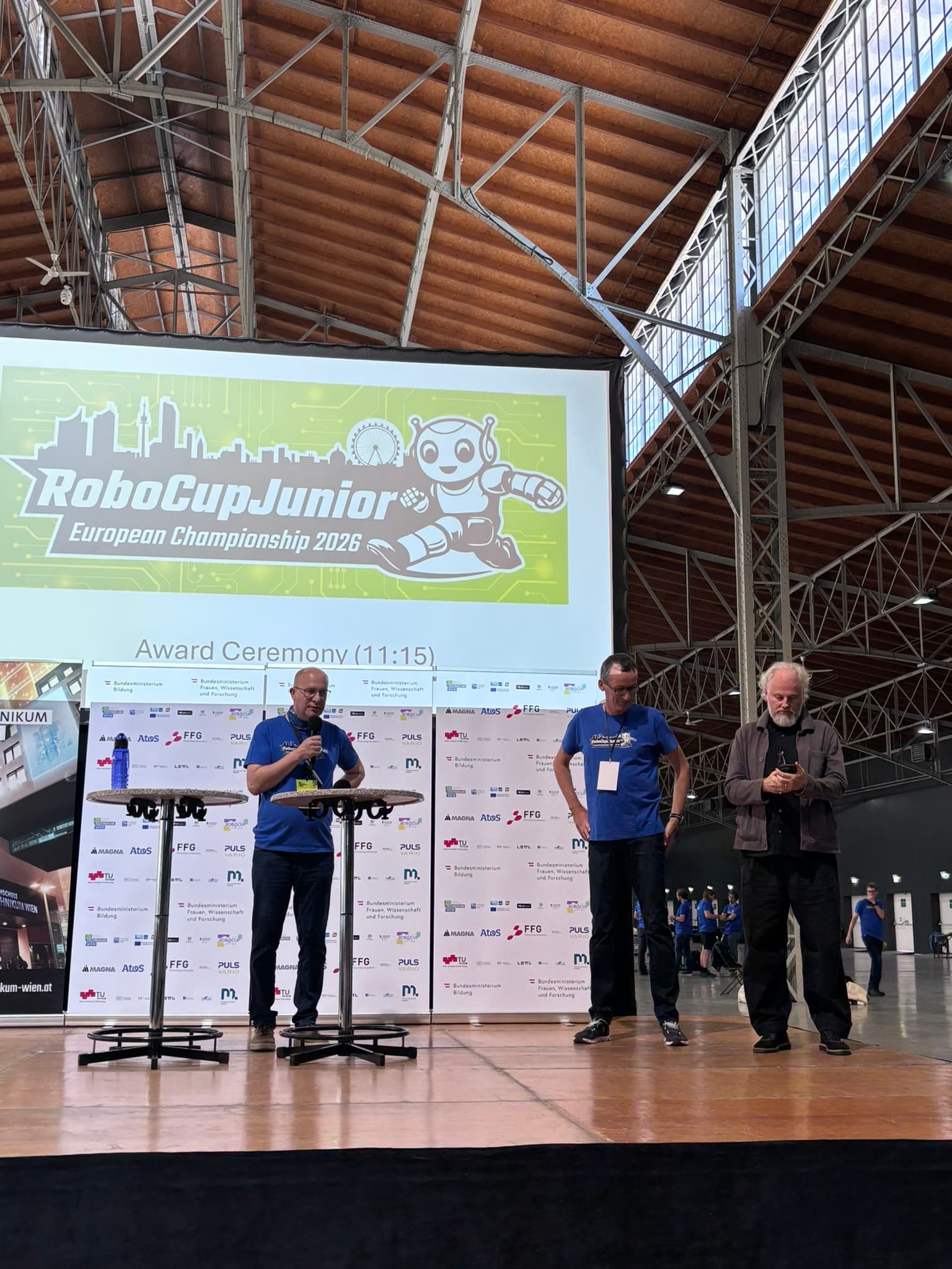 Bei der Siegerehrung der RoboCup Junior-Europameisterschaft 2026 stehen drei Männer auf der Bühne. Hinter ihnen ist in einer geräumigen Halle mit hohen Decken und sichtbaren Metallträgern ein großes Veranstaltungsbanner zu sehen.