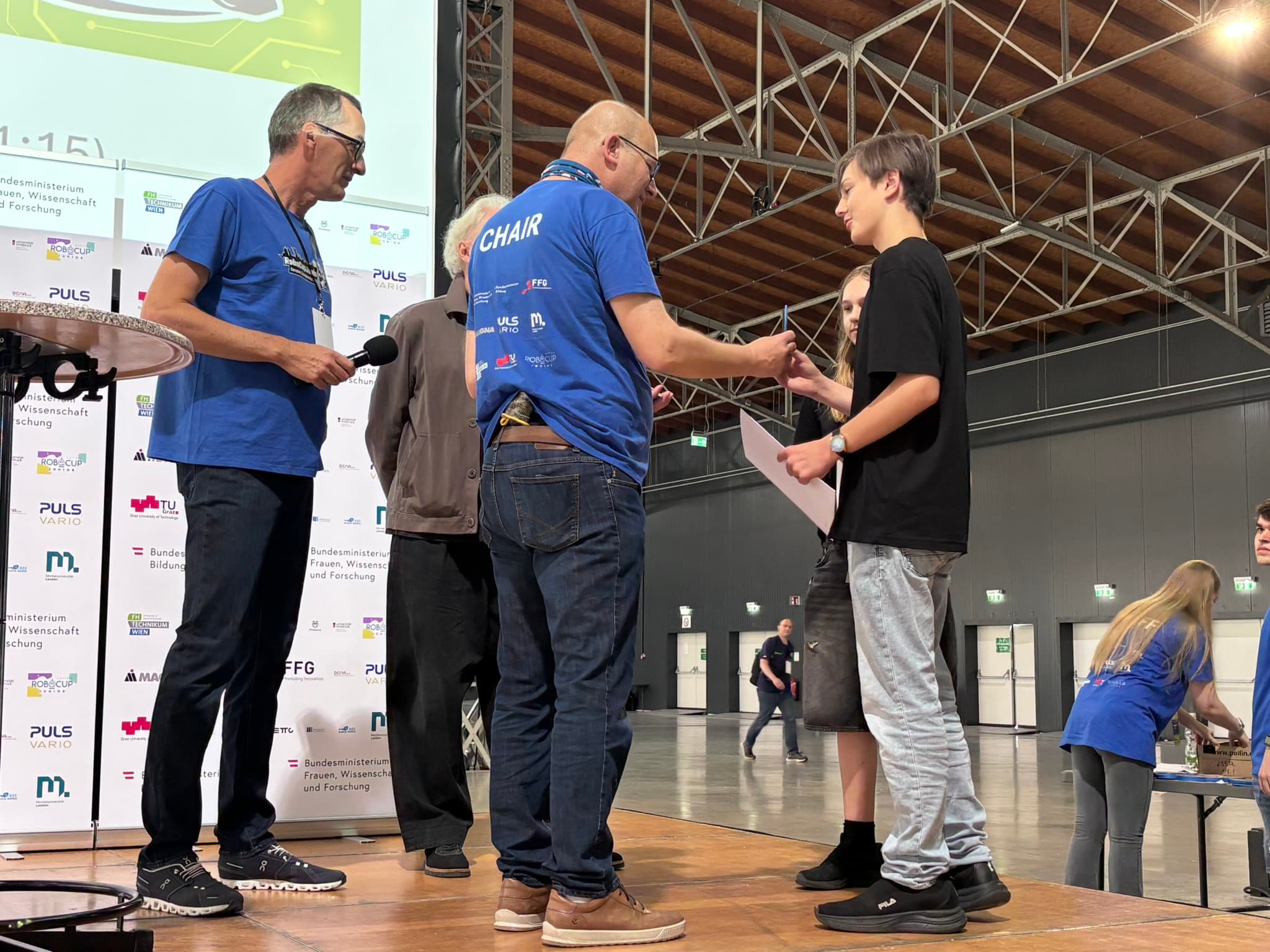 Ein junger Mensch erhält auf einer Bühne bei einer Veranstaltung in einer Halle von einem Erwachsenen in einem blauen CHAIR-T-Shirt eine Urkunde, während andere zuschauen. Im Hintergrund sind Sponsorenbanner und Veranstaltungspersonal zu sehen.
