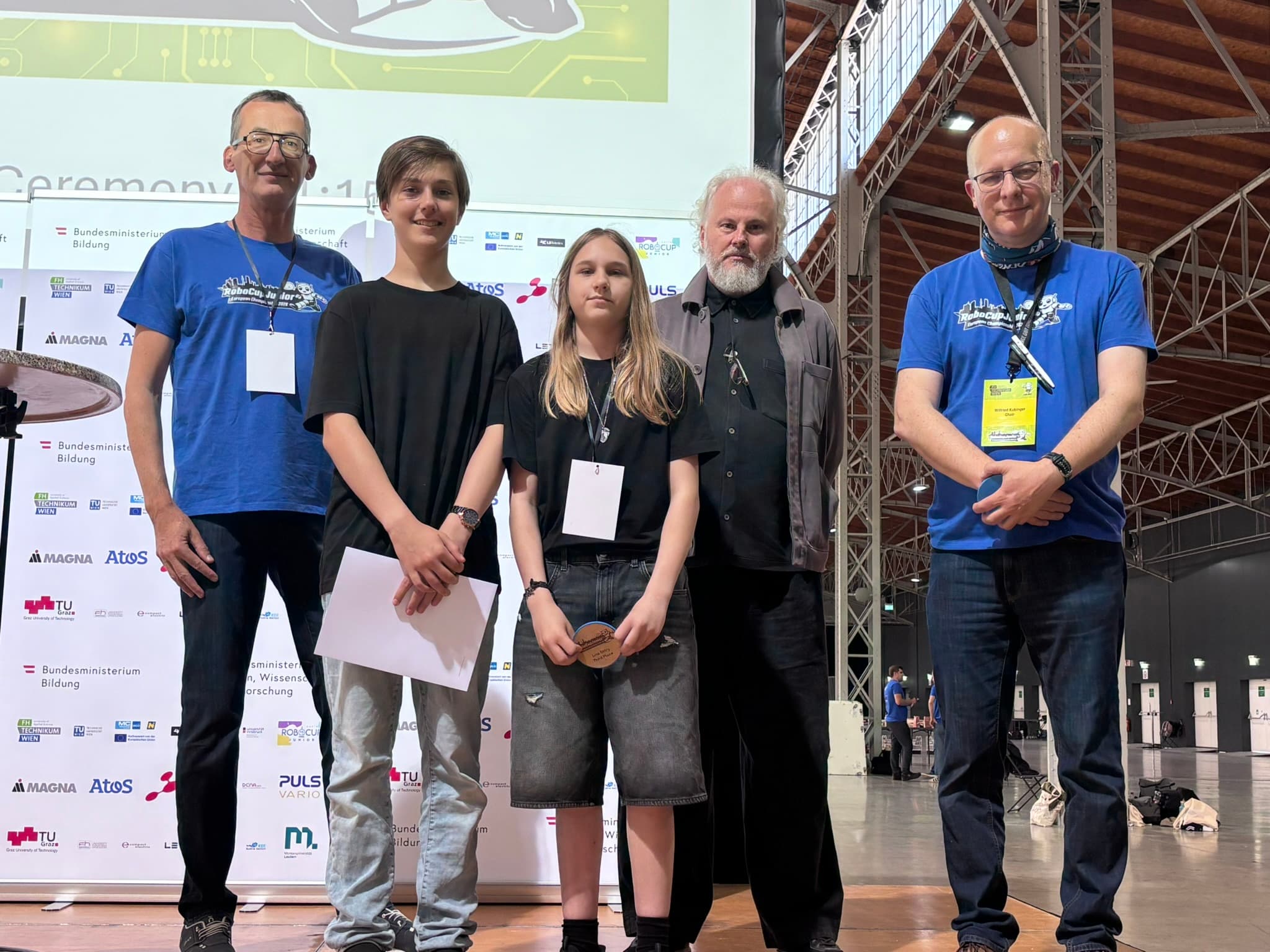 Fünf Personen stehen bei einer Veranstaltung gemeinsam in einem Raum. Auf jeder Seite stehen zwei Erwachsene, die blaue Hemden und Namensschilder tragen, während drei Personen in der Mitte Urkunden und eine Medaille in den Händen halten. Im Hintergrund sind eine Präsentationsleinwand und Banner zu sehen.