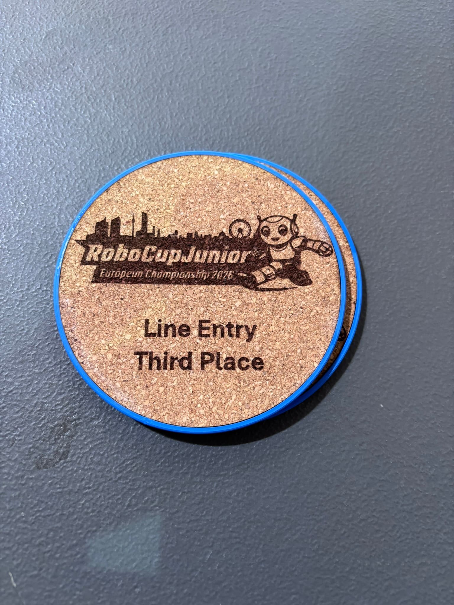 Eine runde Medaille aus Kork mit blauem Rand, auf der „RoboCupJunior-Europameisterschaft 2016, Line Entry, dritter Platz“ zu lesen ist und die mit einer Grafik eines Roboters und einer Stadtsilhouette verziert ist, liegt auf einer strukturierten dunklen Oberfläche.