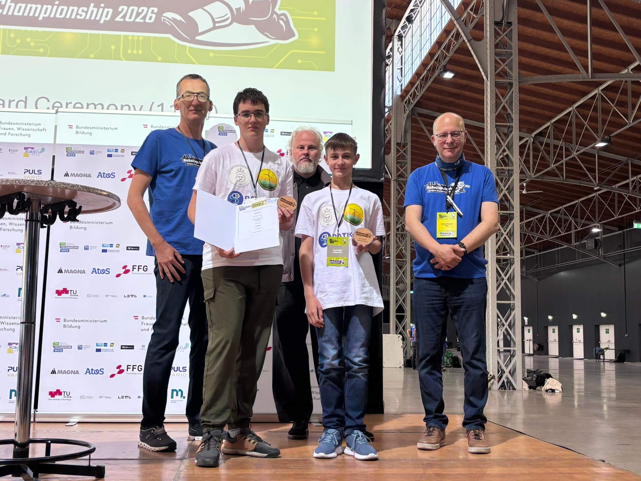 Fünf Personen stehen bei einer Veranstaltung in einer Halle beieinander, einige halten Urkunden und Medaillen in den Händen. Hinter ihnen ist eine große Leinwand zu sehen, auf der „Championship 2026“ und Sponsorenlogos zu sehen sind. Es scheint sich um eine Preisverleihung zu handeln.