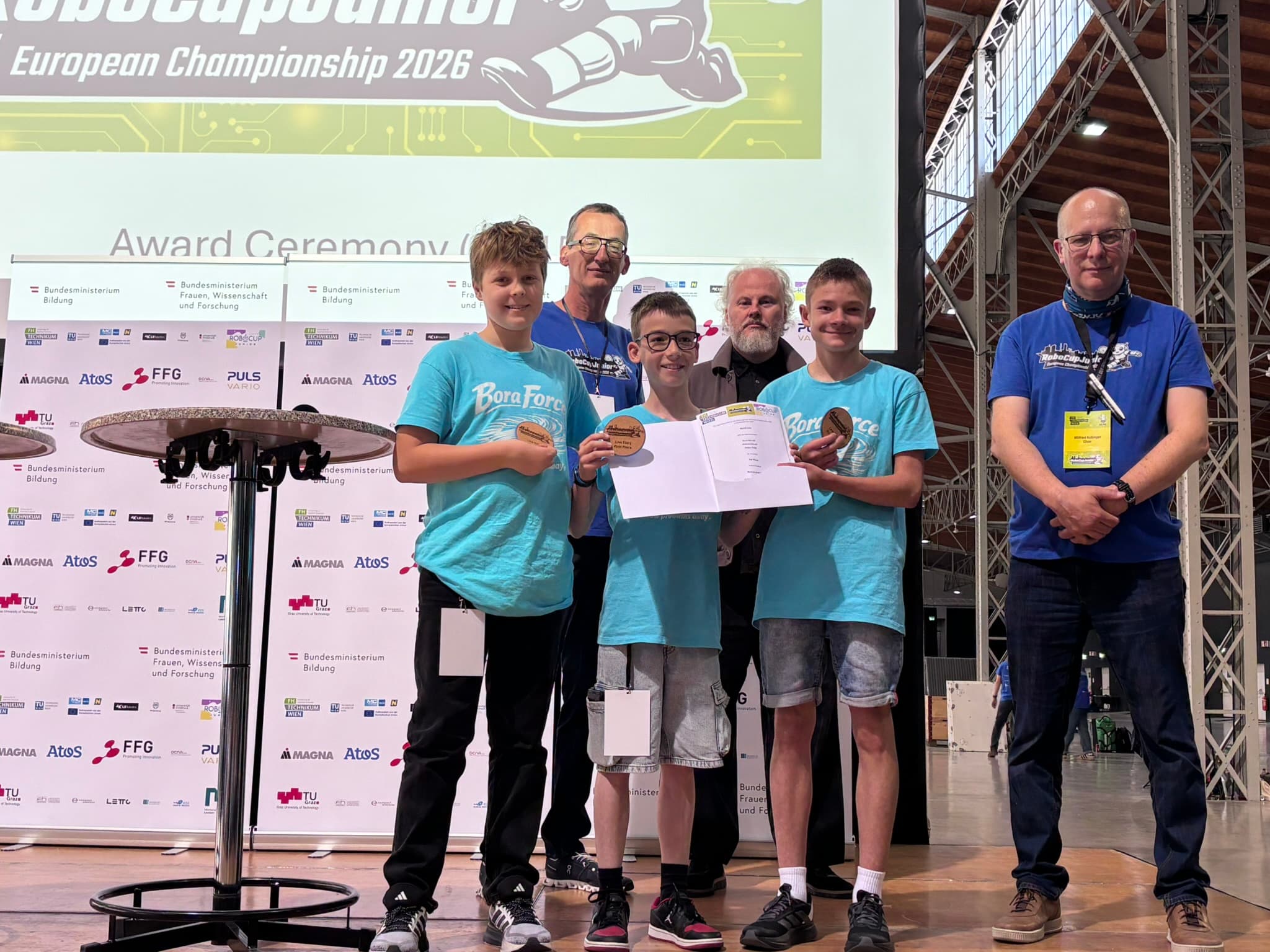 Vier Jungen in einheitlichen blauen T-Shirts halten Medaillen und eine Urkunde in den Händen, während sie zusammen mit drei Erwachsenen bei einer Preisverleihung stehen. Im Hintergrund sind eine große Leinwand und Sponsorenlogos zu sehen.