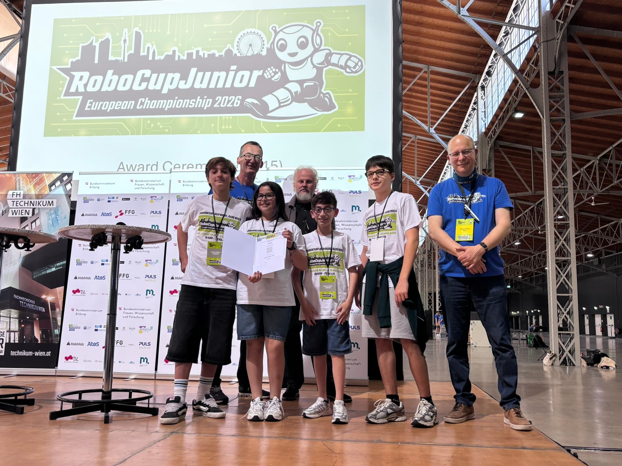 Eine Gruppe von fünf lächelnden Personen – vier Jugendliche und ein Erwachsener – steht bei der RoboCupJunior-Europameisterschaft 2024 auf einer Bühne und hält eine Urkunde in den Händen. Neben ihnen steht ein Mann. Hinter ihnen ist ein großes Veranstaltungsbanner zu sehen.