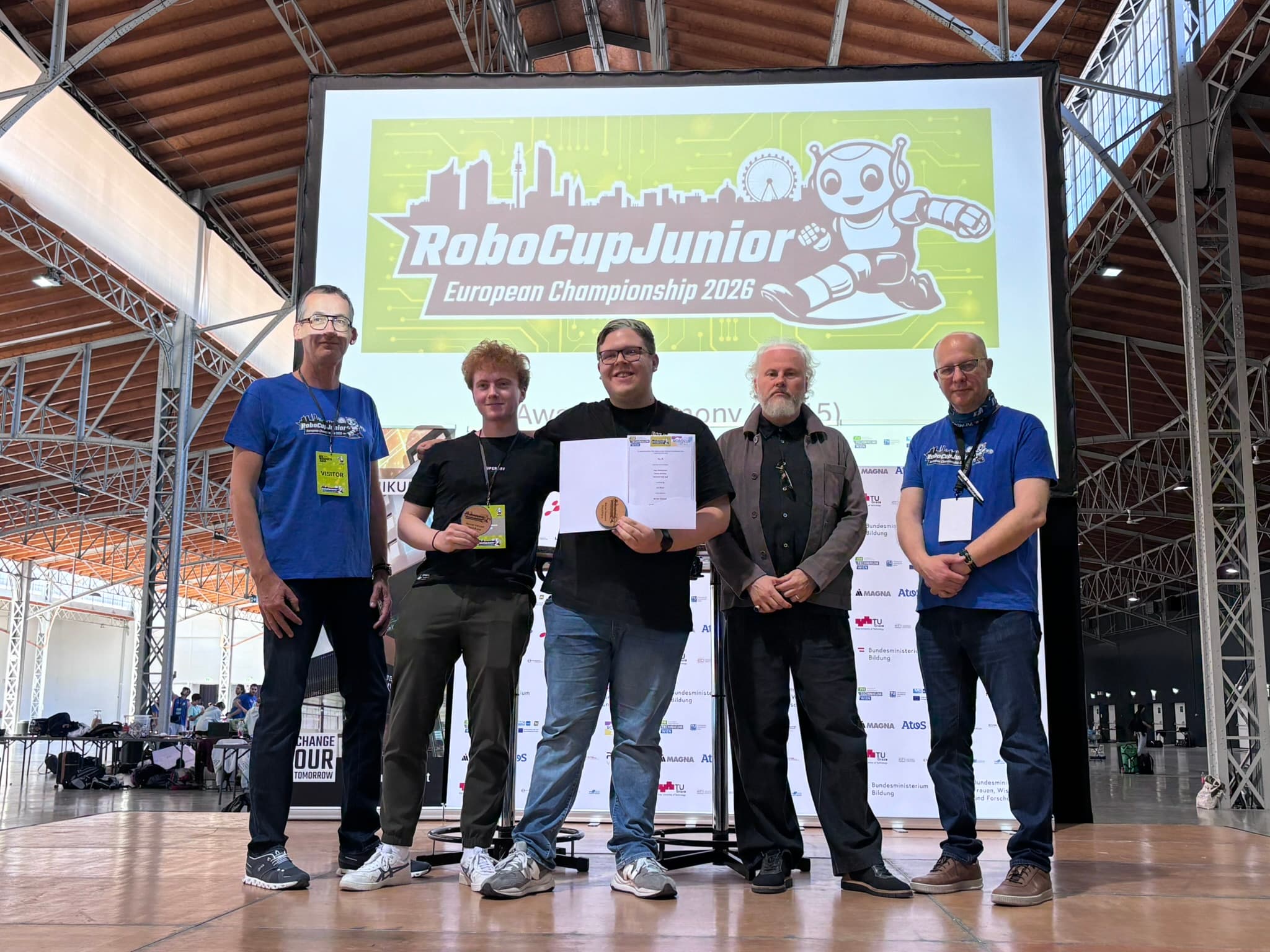 Fünf Personen stehen lächelnd vor einer großen Leinwand, auf der das Logo der RoboCup Junior-Europameisterschaft 2026 zu sehen ist, und halten Urkunden und Auszeichnungen in den Händen – in einem Veranstaltungsraum mit hohen Decken.