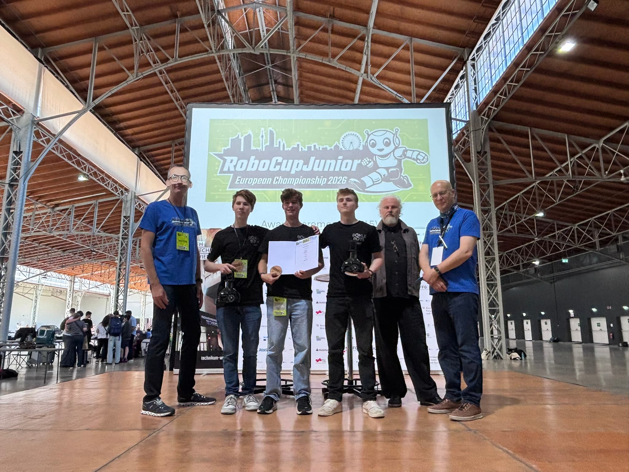 Sechs Personen stehen auf einer Bühne in der Halle und halten Trophäen und Urkunden in den Händen, vor einer großen Leinwand, auf der „RoboCupJunior-Europameisterschaft 2023“ zu lesen ist. Der Schauplatz ist eine geräumige Halle mit hohen Decken und Balken.