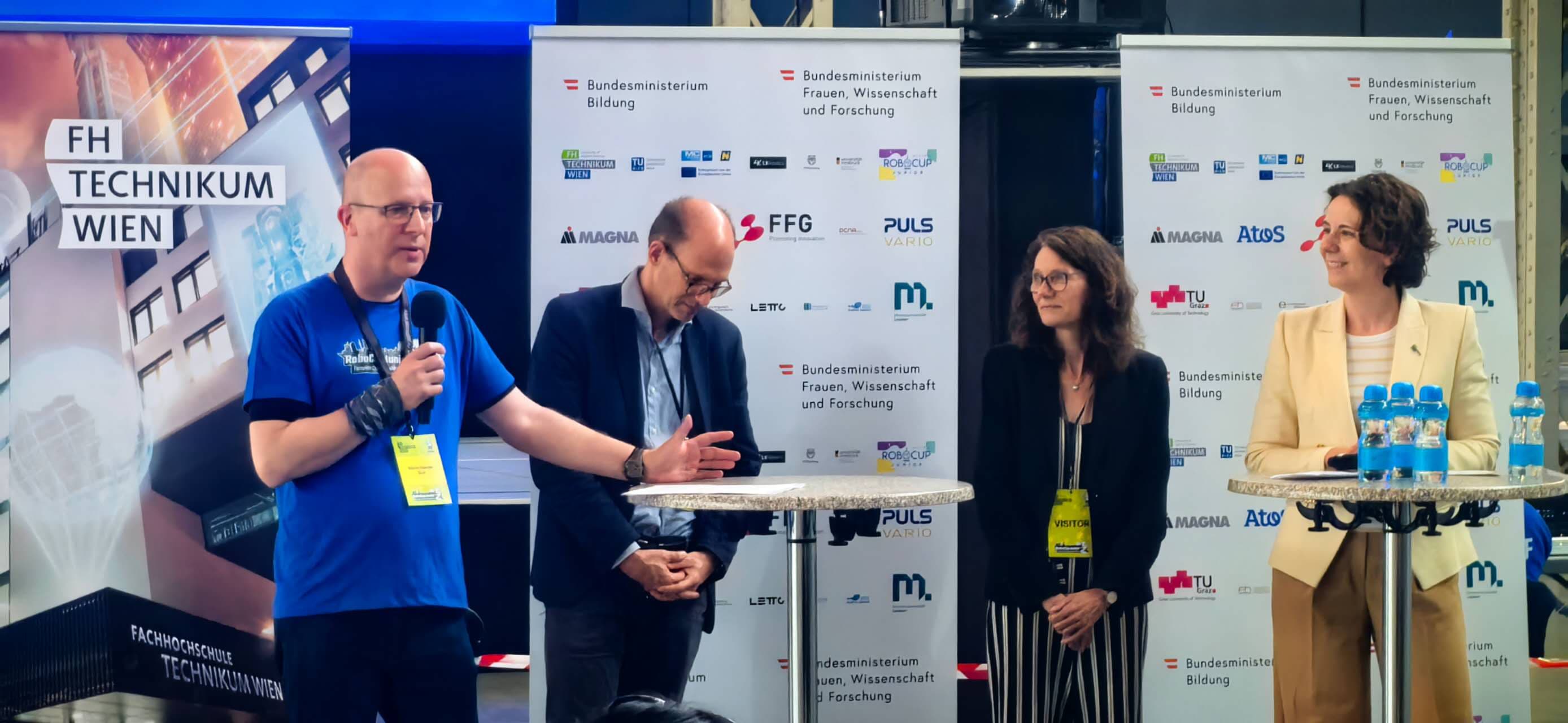Bei einer Veranstaltung an der FH Technikum Wien spricht ein Mann auf einer Bühne in ein Mikrofon, während drei Personen – zwei Frauen und ein Mann – in der Nähe stehen. Im Hintergrund sind Banner und ein Tisch mit Wasserflaschen zu sehen.