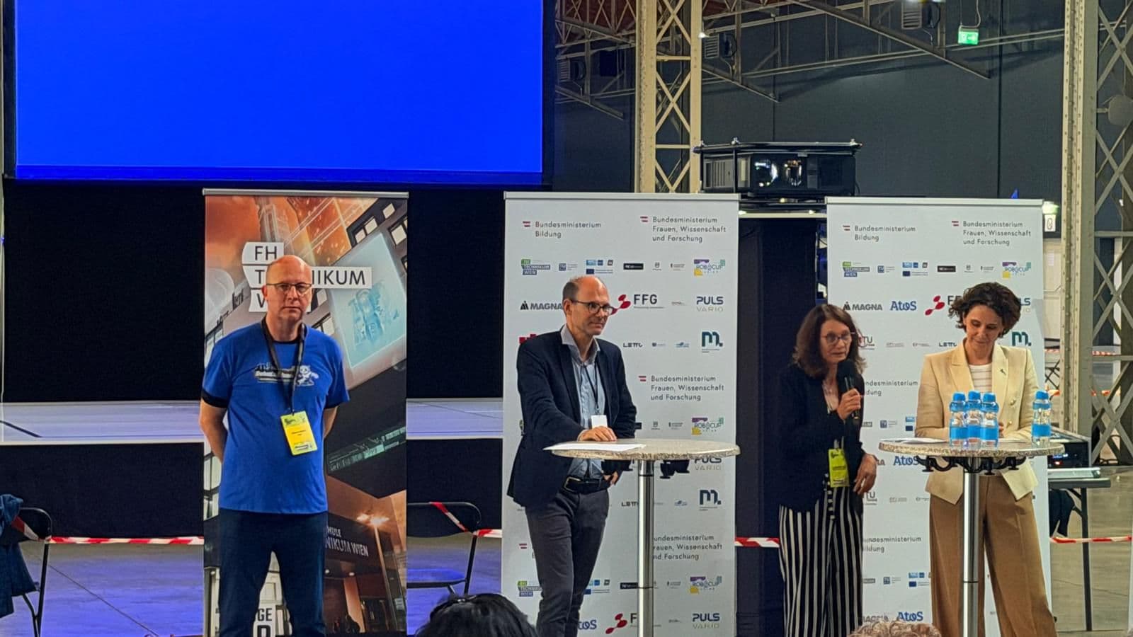 Vier Personen stehen auf der Bühne einer Technologieveranstaltung; drei sitzen an einem Tisch mit Mikrofonen und Wasserflaschen, eine steht daneben. Im Hintergrund sind ein blauer Hintergrund und Banner mit Logos zu sehen.
