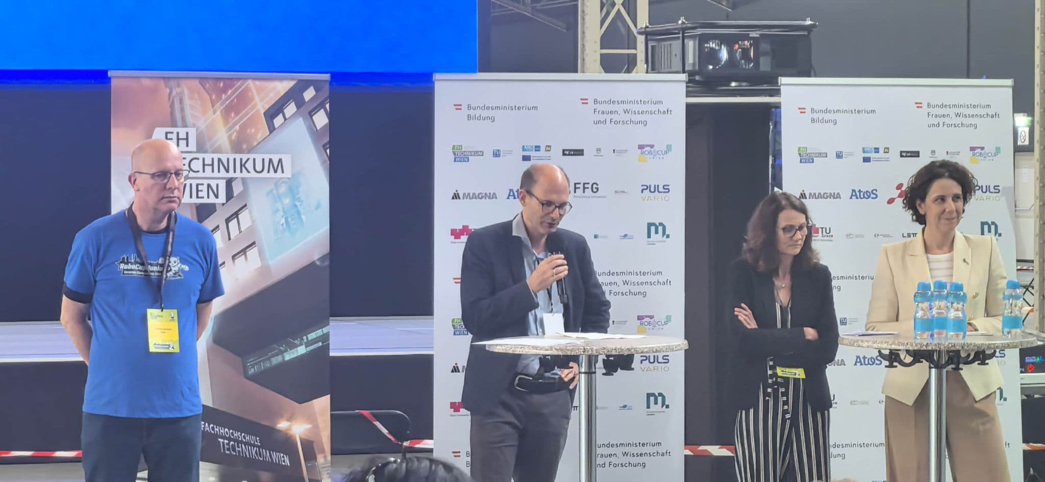 Vier Personen stehen auf einer Bühne bei einer Technologieveranstaltung, hinter ihnen sind zwei Banner mit Logos und Text zu sehen. Ein Mann spricht in ein Mikrofon, während die anderen zuhören. Im Hintergrund ist ein großer blauer Bildschirm zu sehen.
