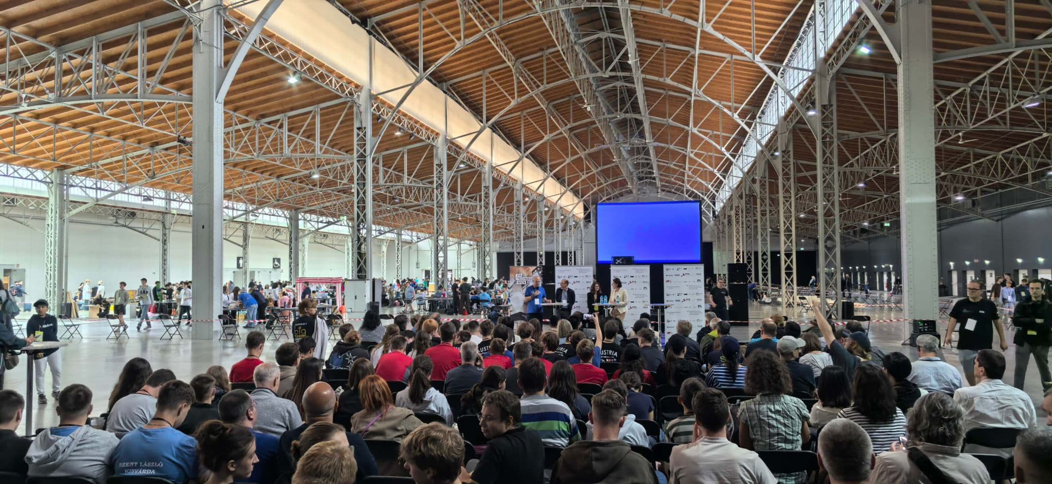 In einer geräumigen Industriehalle mit hoher Decke und freiliegenden Balken sitzt eine große Gruppe von Menschen mit Blick auf eine Bühne. Eine große Leinwand im vorderen Bereich ist blau beleuchtet, und rund um die Halle sind verschiedene Stände und Geräte aufgestellt.