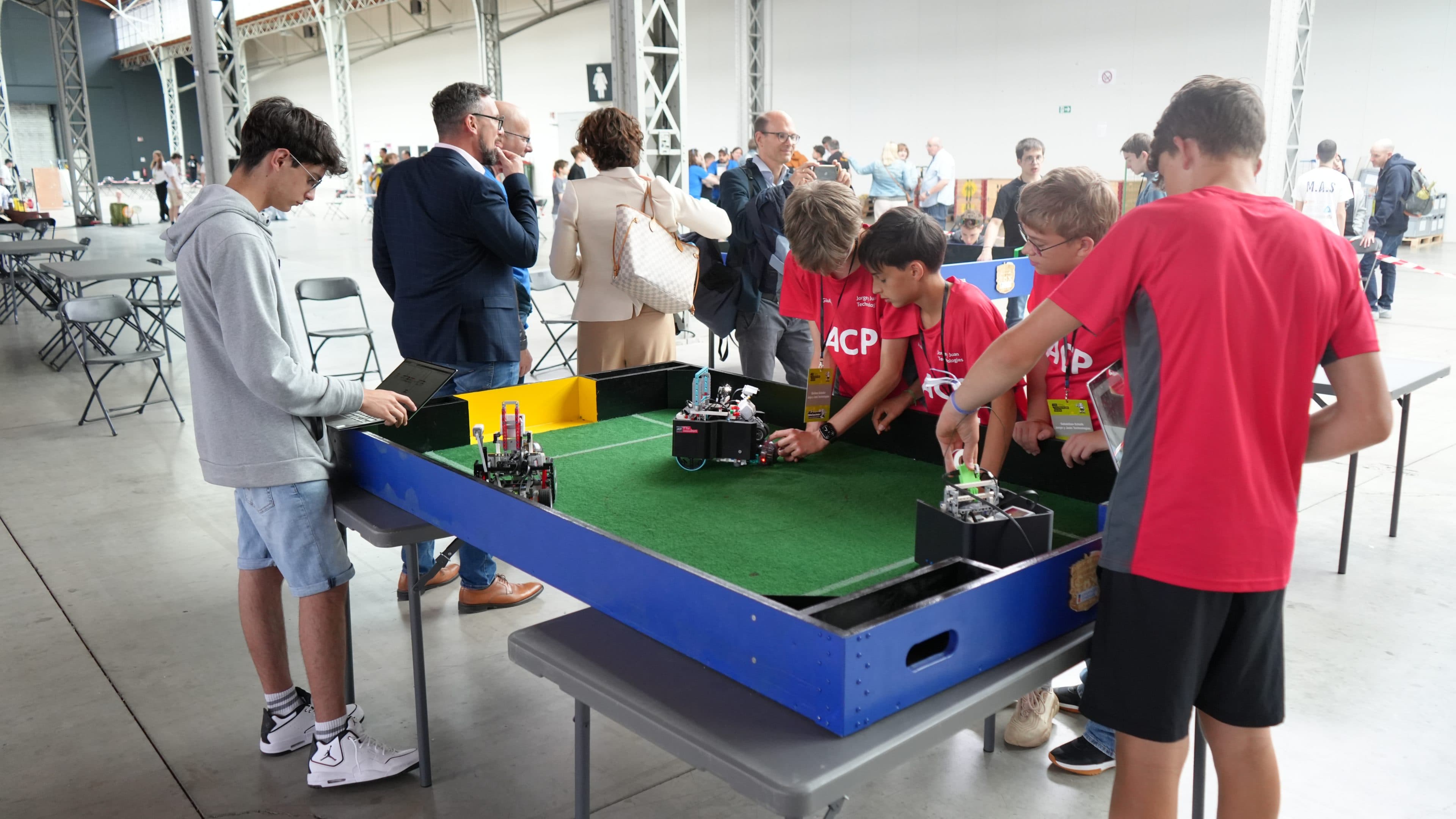 Eine Gruppe von Jungen steuert kleine Roboter auf einem grünen Tisch, der einem Miniaturfußballfeld ähnelt, während mehrere Erwachsene in einem geräumigen Veranstaltungsraum zuschauen.