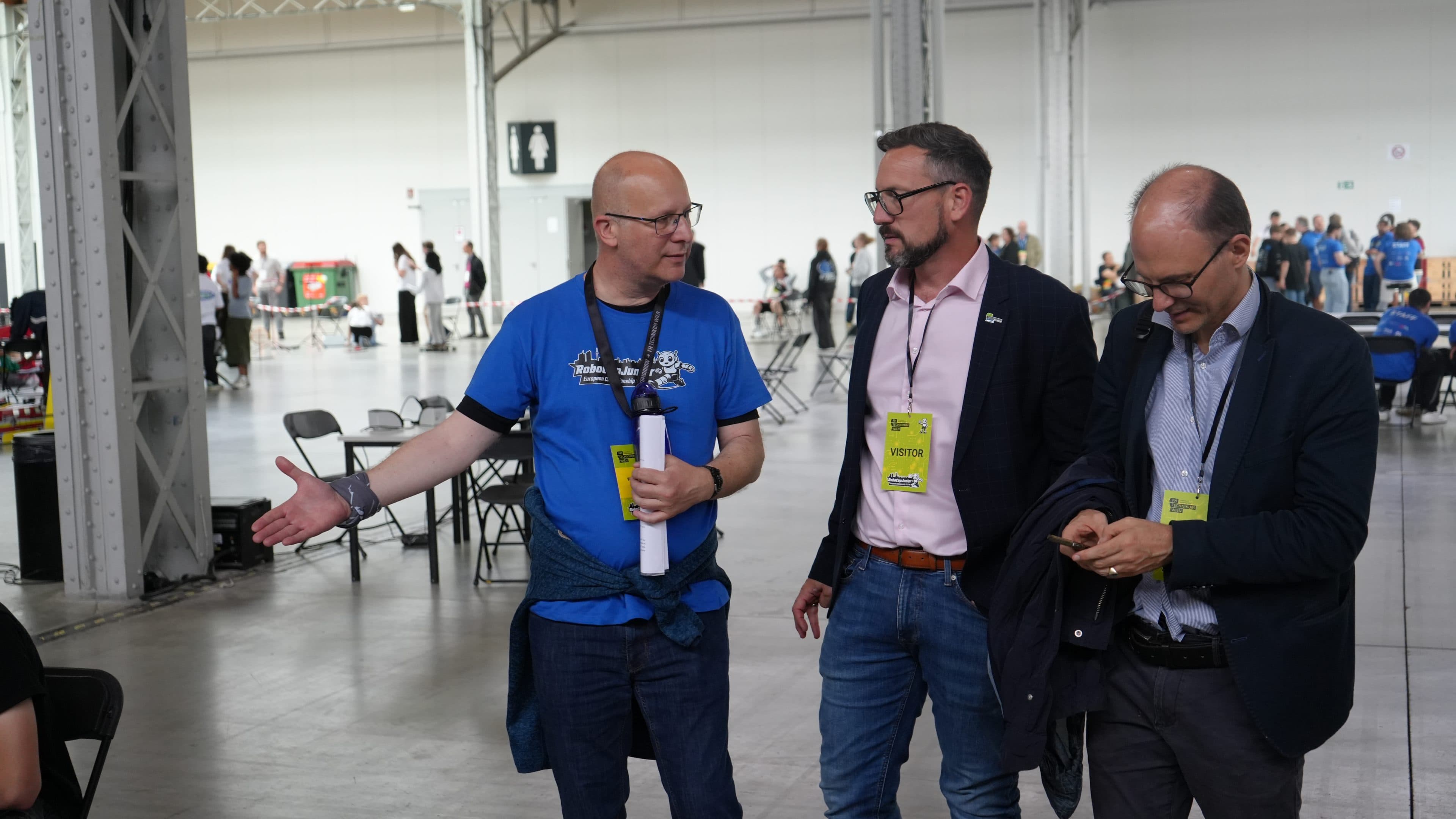 Drei Männer mit Konferenzausweisen unterhalten sich in einem großen Veranstaltungsraum. Ein Mann gestikuliert mit der Hand, ein anderer hört zu, und der dritte schaut auf sein Handy. Im Hintergrund sind weitere Personen und Stühle zu sehen.