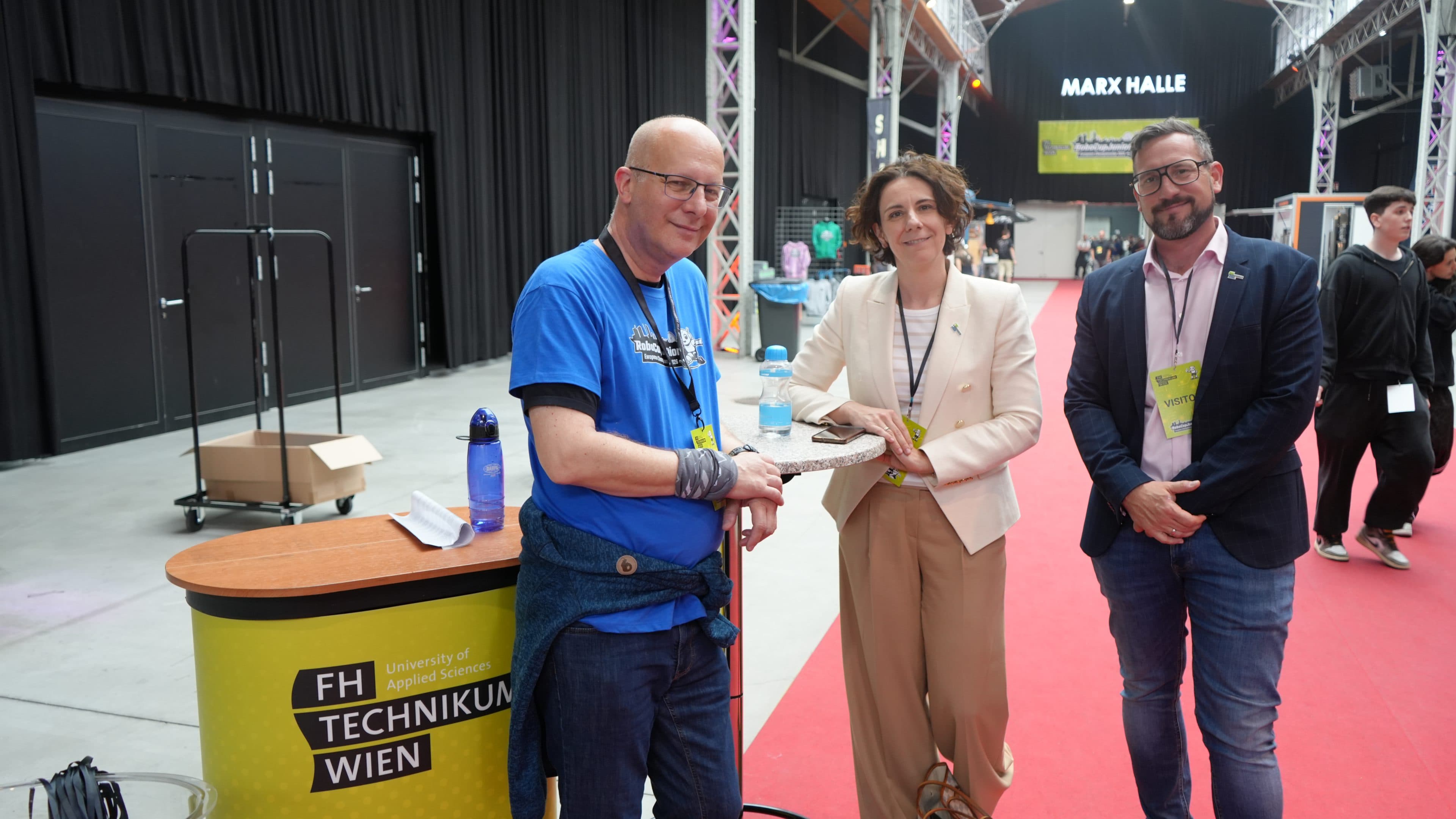 Drei Personen stehen lächelnd neben einem Stand mit der Aufschrift „FH Technikum Wien“ auf einer Indoor-Veranstaltung. Die Person links trägt ein blaues Hemd, während die beiden anderen Business-Kleidung tragen und auf einem roten Teppich stehen.