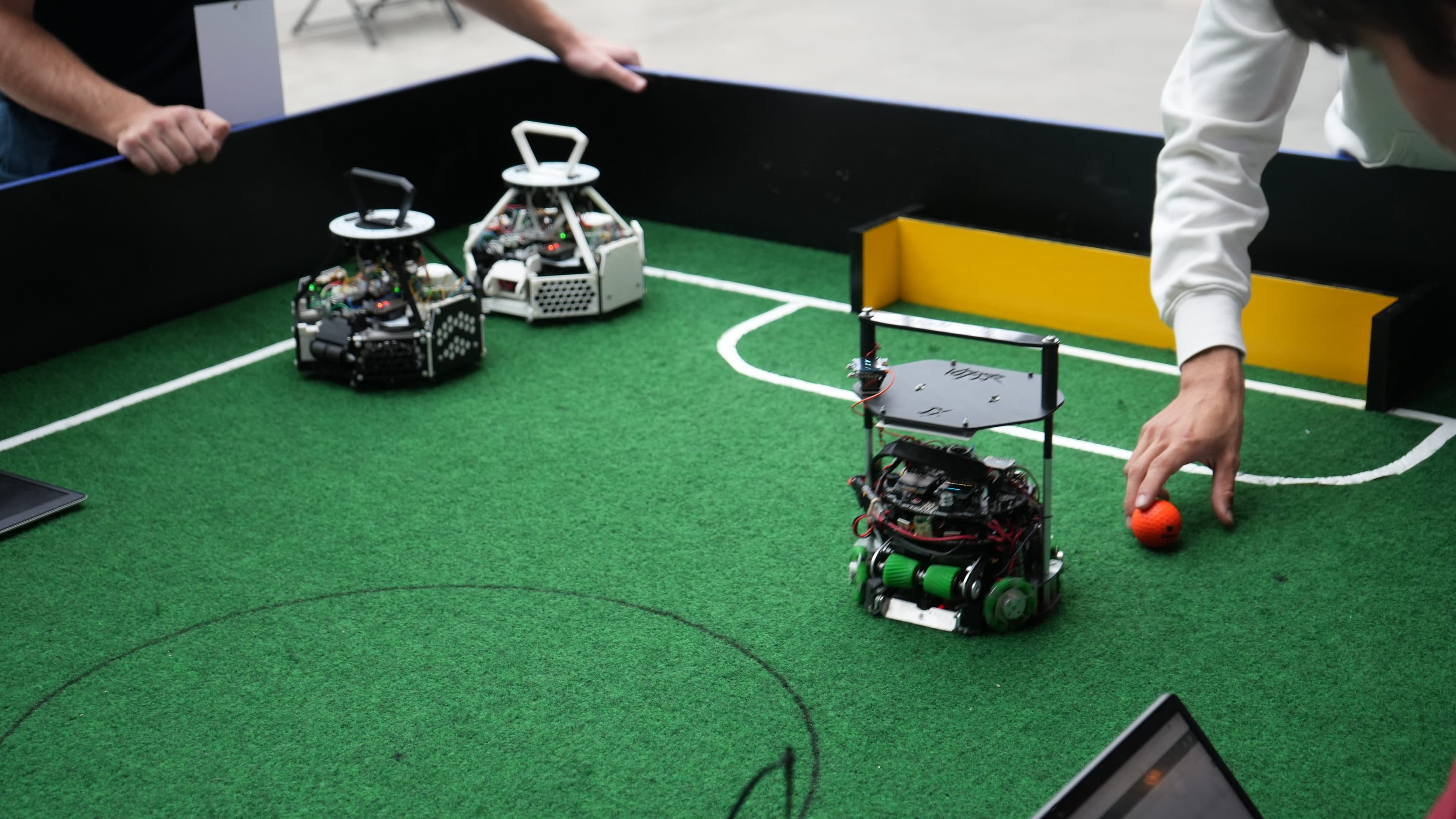Drei kleine Roboter stehen auf einem grünen Feld, das einem Fußballplatz ähnelt. Zwei Personen nehmen Einstellungen an den Robotern vor, und eine Person legt einen orangefarbenen Ball in der Nähe des Torraums ab. Die Szene scheint ein Roboterfußballspiel zu sein.