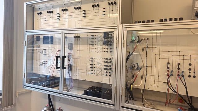 Ein elektrischer Schaltschrank für ein Labor mit Schaltplänen, Schaltern, Drehknöpfen und Kabeln hinter transparenten Glastüren. Unterhalb des Schaltschranks sind Geräte und Kabel zu sehen, und die Anlage wirkt übersichtlich.