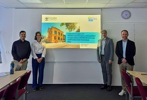 Vier Personen stehen in einem Klassenzimmer vor einer Präsentationswand. Auf der Wand ist eine Folie mit Text und dem Bild eines Gebäudes zu sehen. Tische, Stühle und ein an der Decke montierter Projektor sind zu erkennen.