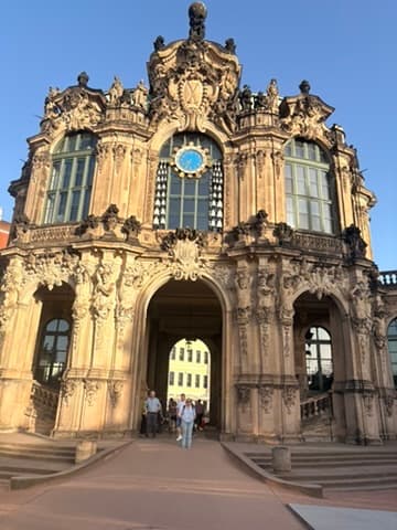 Ein prunkvolles Gebäude im Barockstil mit einem großen Torbogen als Eingang, aufwendigen Steinmetzarbeiten, hohen Fenstern und einer blauen Uhr über dem Torbogen. An einem sonnigen Tag gehen mehrere Menschen durch den Eingang.