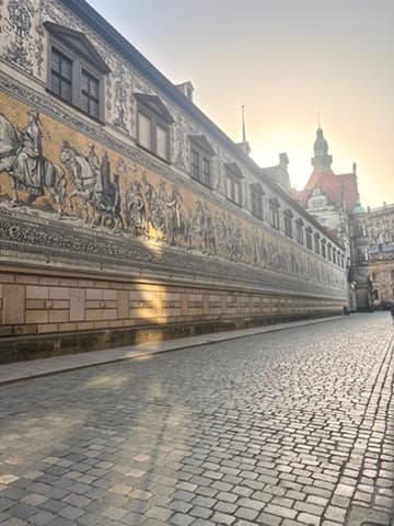 Eine lange Kopfsteinpflasterstraße verläuft entlang eines historischen Gebäudes, das mit einem großen Wandgemälde geschmückt ist, auf dem Menschen zu Pferd dargestellt sind; ein Teil der Wand wird vom Sonnenlicht beleuchtet. Im Hintergrund ragt ein kunstvoll verzierter Turm empor.