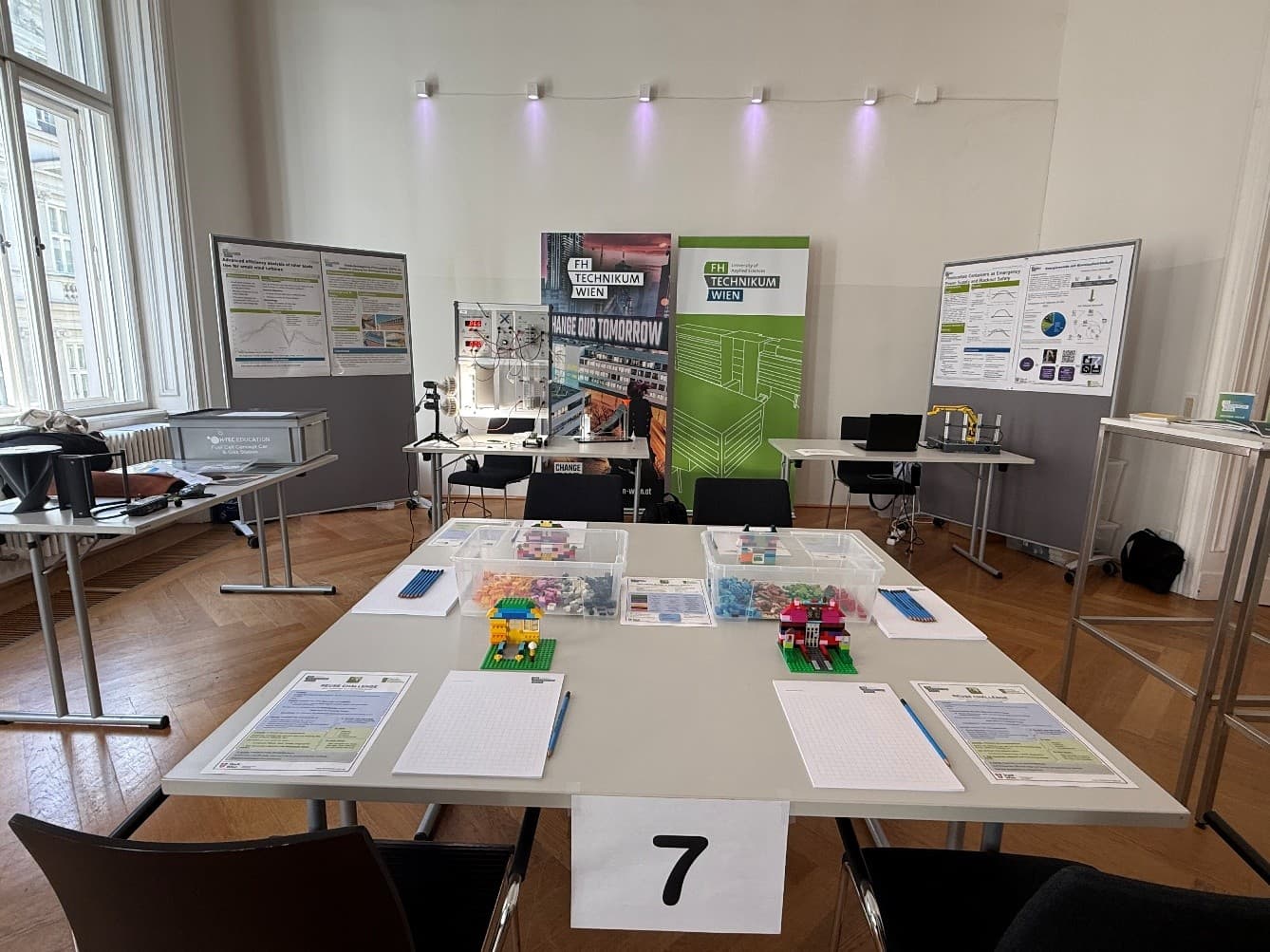 Ein Klassenzimmer mit Tischen, Stühlen und bunten LEGO-Modellen auf einem zentralen Tisch mit der Nummer 7, umgeben von Plakaten, Diagrammen und Informationstafeln zum Thema Technik an den Wänden.