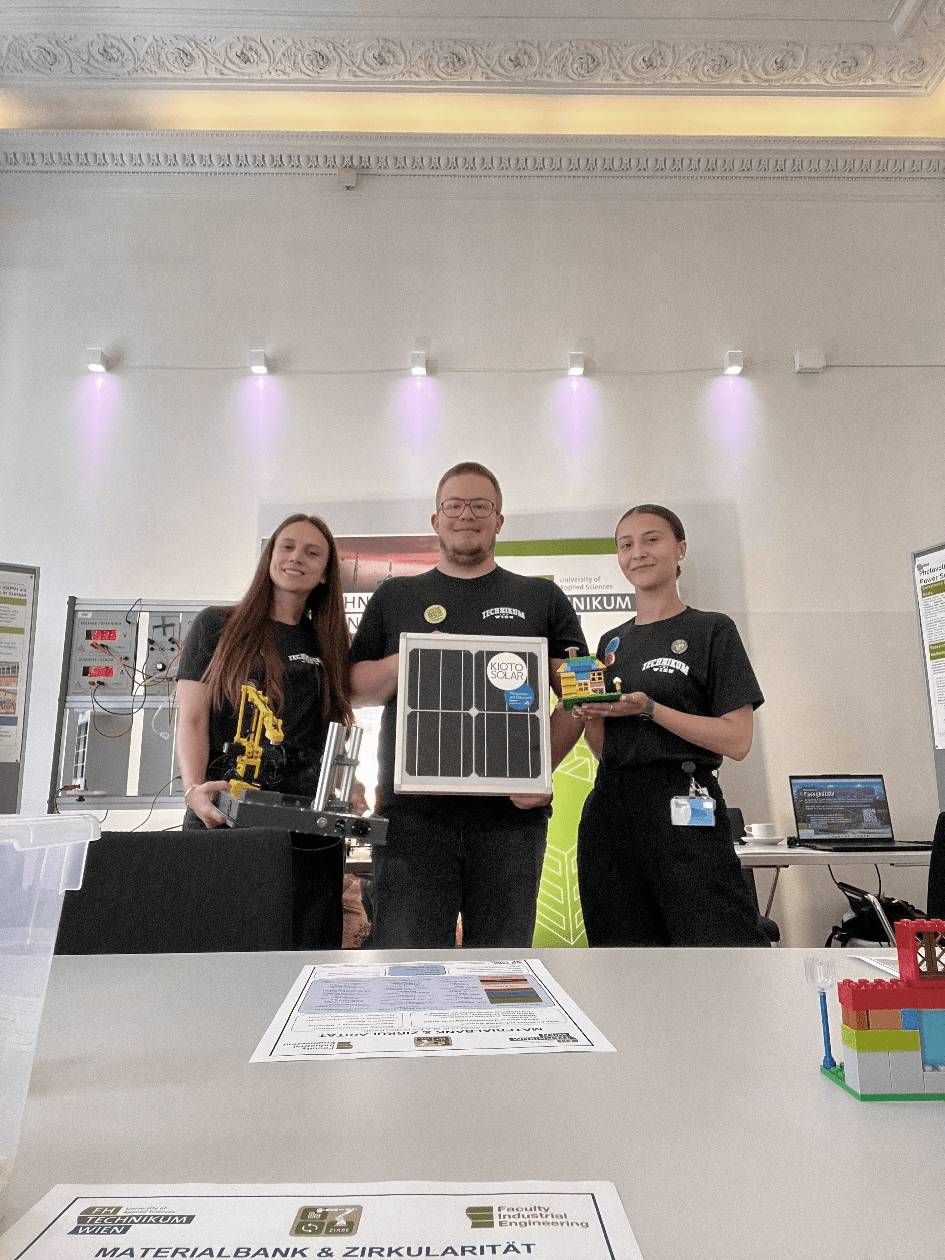 Vier Personen stehen hinter einem Tisch, auf dem ein Solarpanel-Projekt und Informationsmaterial ausgestellt sind; einige tragen einheitliche T-Shirts. Der Schauplatz scheint eine Ausstellung in einem Innenraum oder eine Wissenschaftsmesse zu sein, im Hintergrund sind Plakate und Ausstellungsstücke zu sehen.