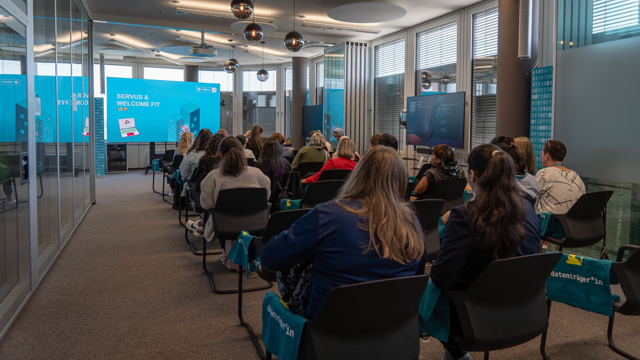 Eine Gruppe von Menschen sitzt in einem modernen Konferenzraum vor einer großen Leinwand, auf der während einer Präsentation „Servus & Welcome Fit“ zu sehen ist. Der Raum hat Glaswände und ist in Blau gehalten; die Teilnehmer tragen blaue Tragetaschen.