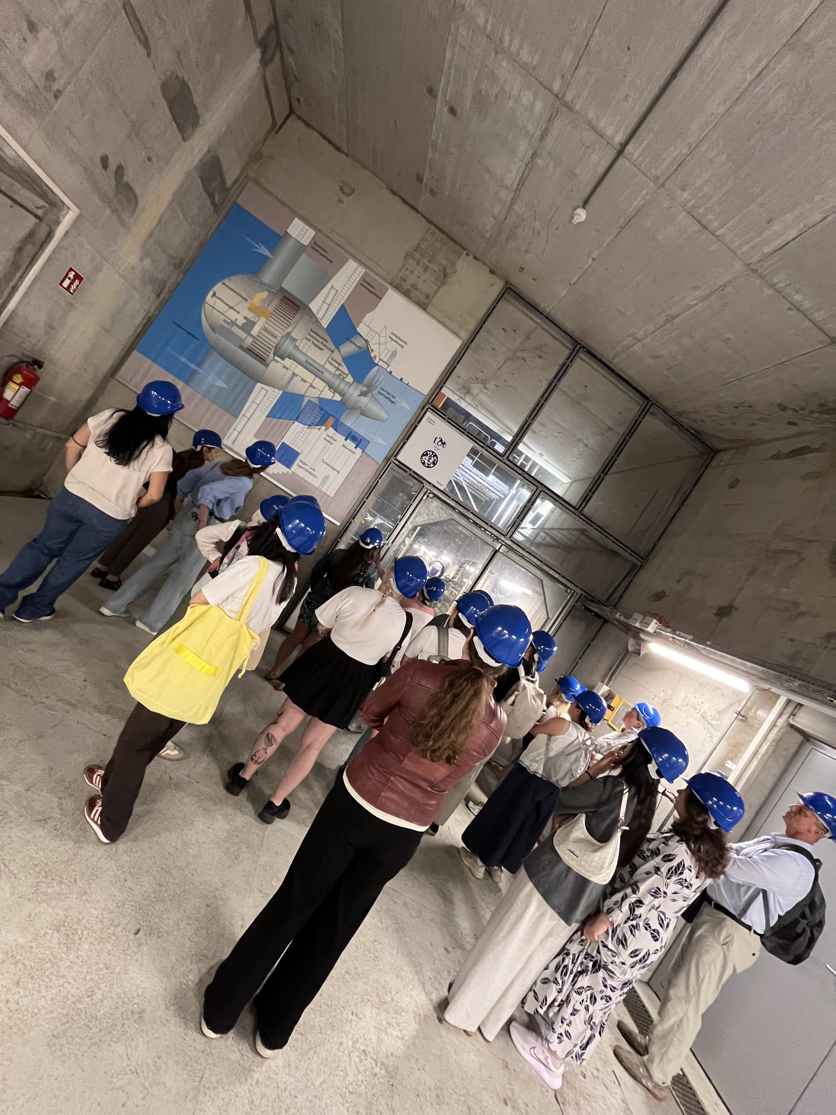 Eine Gruppe von Menschen mit blauen Schutzhelmen steht in einem Betonraum und blickt auf eine große Grafik an der Wand. Die meisten stehen mit dem Rücken zur Kamera, und eine Person ganz vorne trägt eine gelbe Tasche.