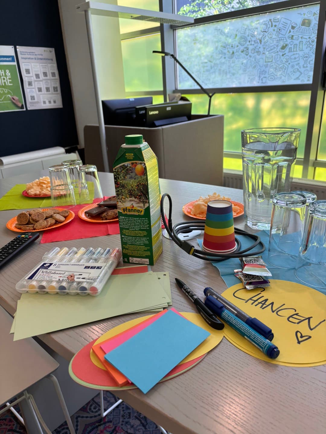 Ein für ein Treffen gedeckter Tisch mit Tellern voller Snacks, einem Karton Saft, Wasserkrügen, Gläsern, bunten Filzstiften, Haftnotizen, einem Stapel Pappbecher und einem gelben Zettel mit der Aufschrift „CHANCEN“ und einem Herz.