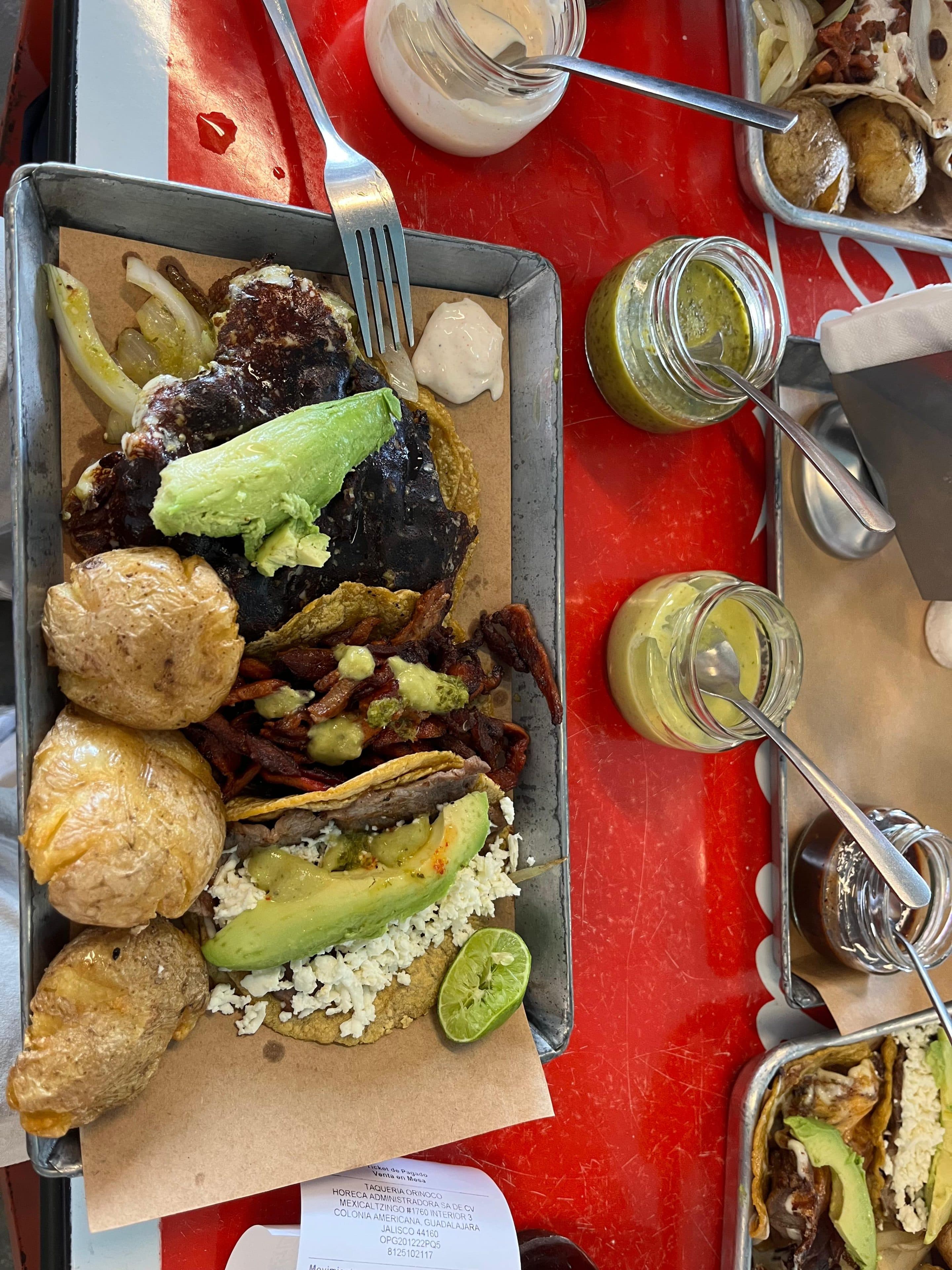 Auf einem Metalltablett liegen ein gegrilltes Steak mit Avocado, Bratkartoffeln, Tacos mit Käse und Avocado sowie eine Limettenscheibe. Auf einem roten Tisch daneben stehen Gläser mit grüner und gelber Soße. Auf dem Tablett liegt eine Gabel.