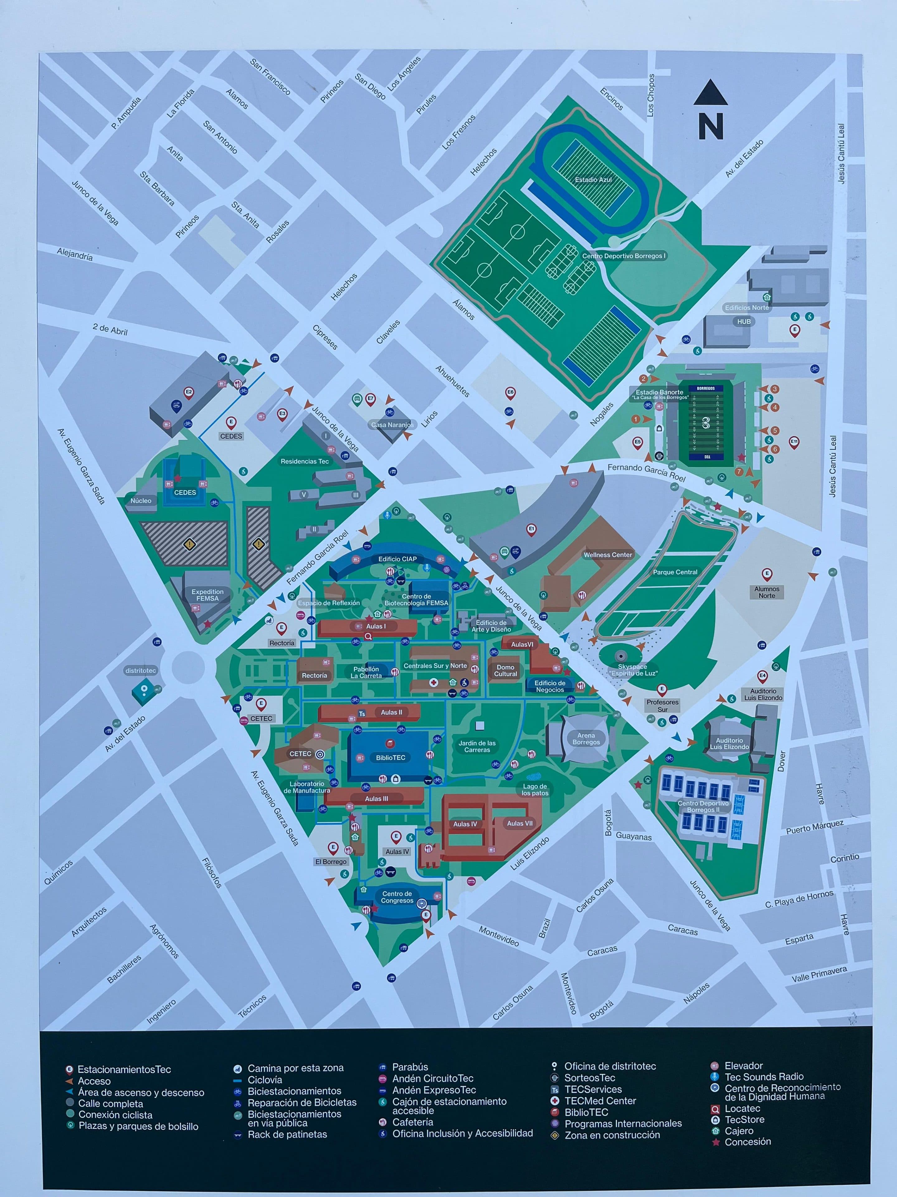 Karte eines Universitätsgeländes mit Gebäuden, Wegen, Sportplätzen und beschrifteten Bereichen, umgeben von Stadtstraßen. Symbole kennzeichnen verschiedene Einrichtungen, und eine Legende am unteren Rand erläutert die Kartensymbole. Der Norden ist oben rechts angegeben.