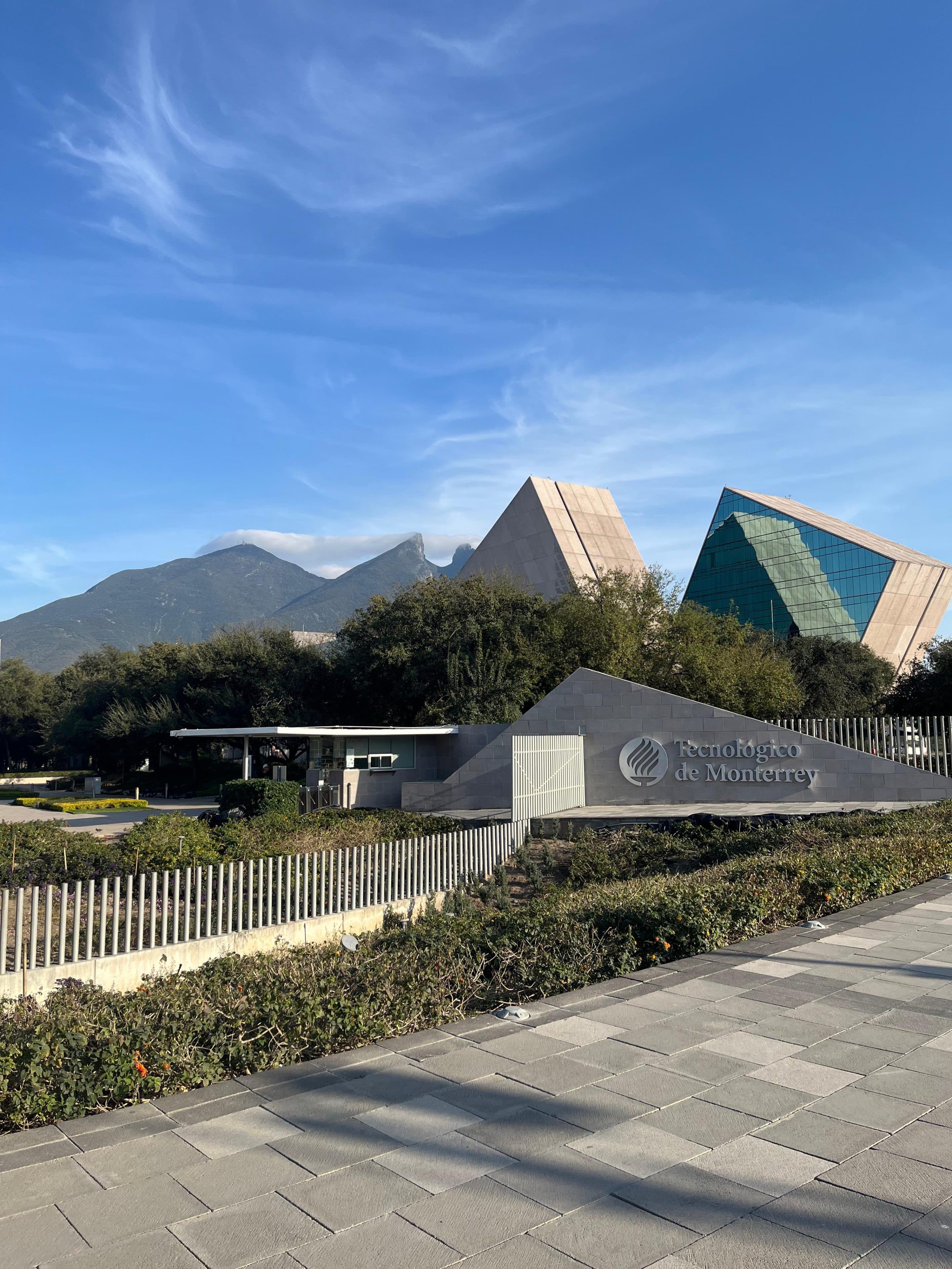 Moderne Gebäude mit dreieckigen Glasfassaden am Tecnológico de Monterrey, vor dem Hintergrund von Bäumen und Bergen unter einem blauen Himmel mit vereinzelten Wolken. Im Vordergrund sind auf einem Steinschild ein Logo und ein Name zu sehen.