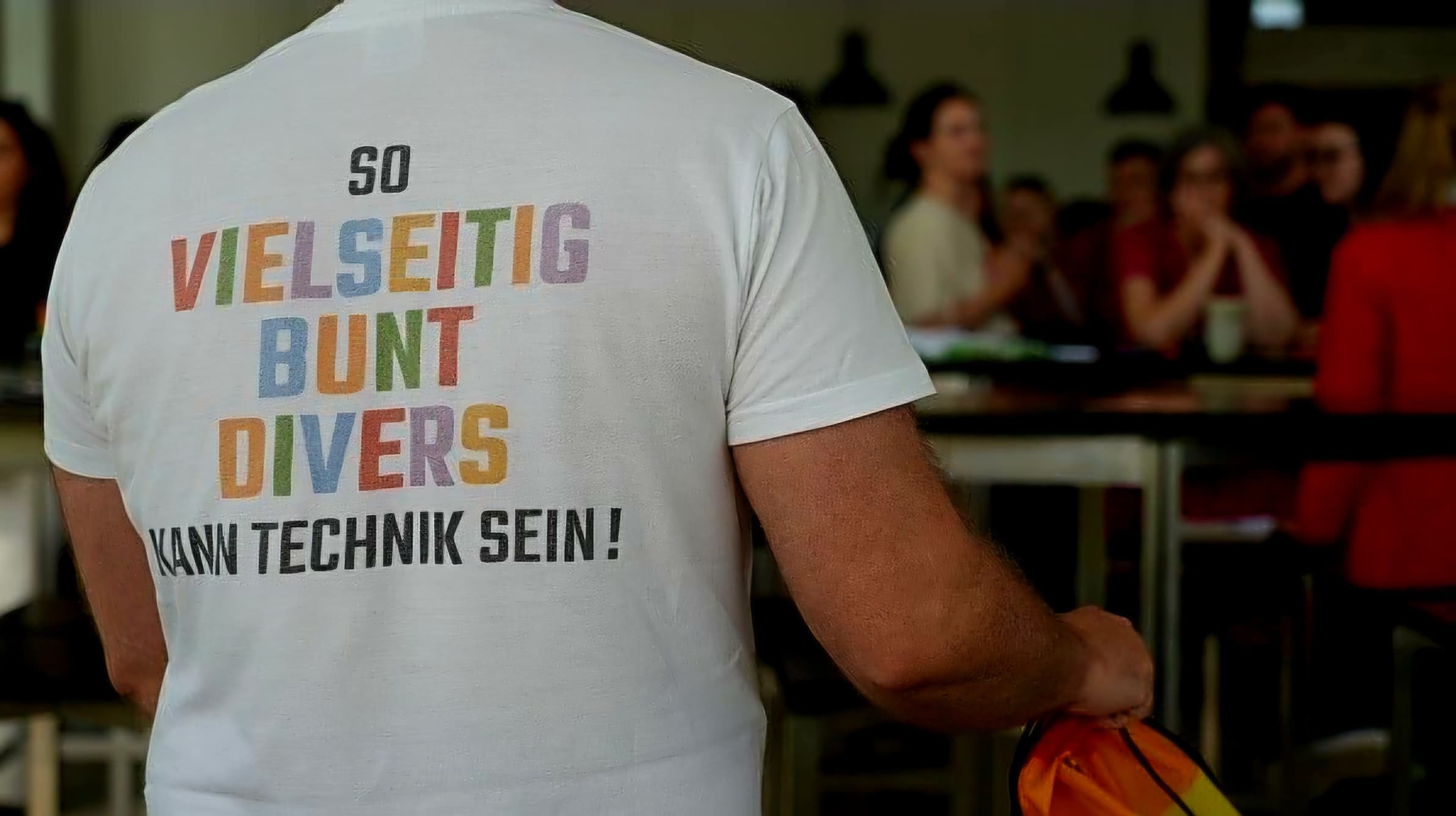 Eine Person, die ein weißes T-Shirt mit buntem deutschen Schriftzug trägt: „So vielseitig, bunt, divers kann Technik sein!“ Im Hintergrund sind sitzende Personen zu sehen, die unscharf dargestellt sind, was auf eine gesellschaftliche oder berufliche Zusammenkunft hindeutet.