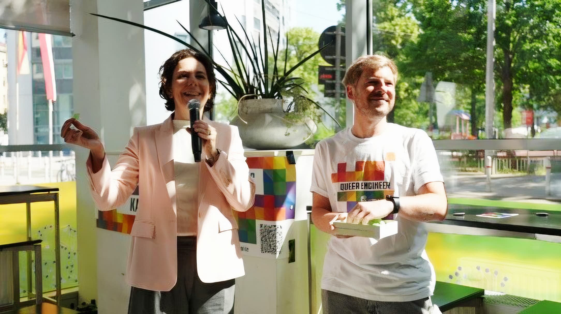 Zwei Personen stehen in einem Raum vor großen Fenstern. Die eine trägt einen hellrosa Blazer, hält ein Mikrofon in der Hand und gestikuliert; die andere, die ein weißes „QUEER HIGHENER“-T-Shirt trägt, lächelt und hält ein Buch in der Hand. Sonnenlicht strömt herein, und im Hintergrund sind Regenbogen-Dekorationen zu sehen.